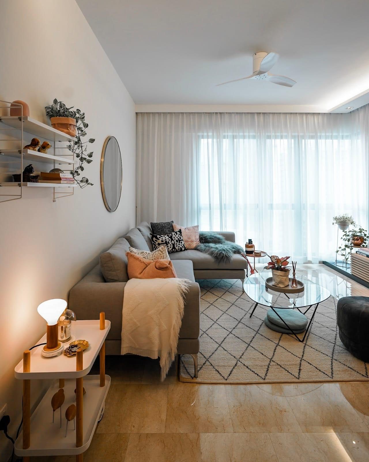 A living room in a Newton condominium with a grey sectional under sheer curtains drawn across the full window wall, a round mirror and wire shelves on the wall opposite, and a glass table on a diamond-patterned rug.