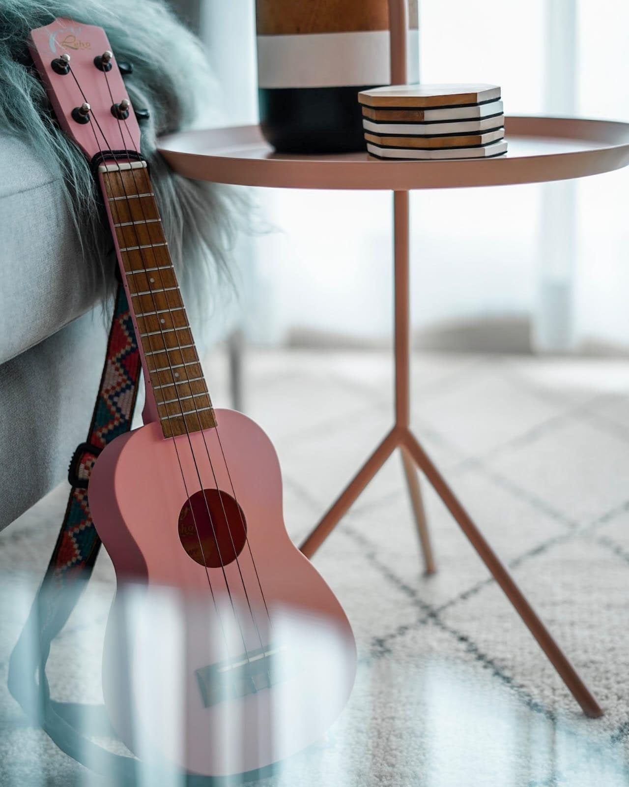 A pink ukulele on a woven strap leaning against a grey sofa in a Newton condominium, beside a blush side table holding a stack of stone coasters.