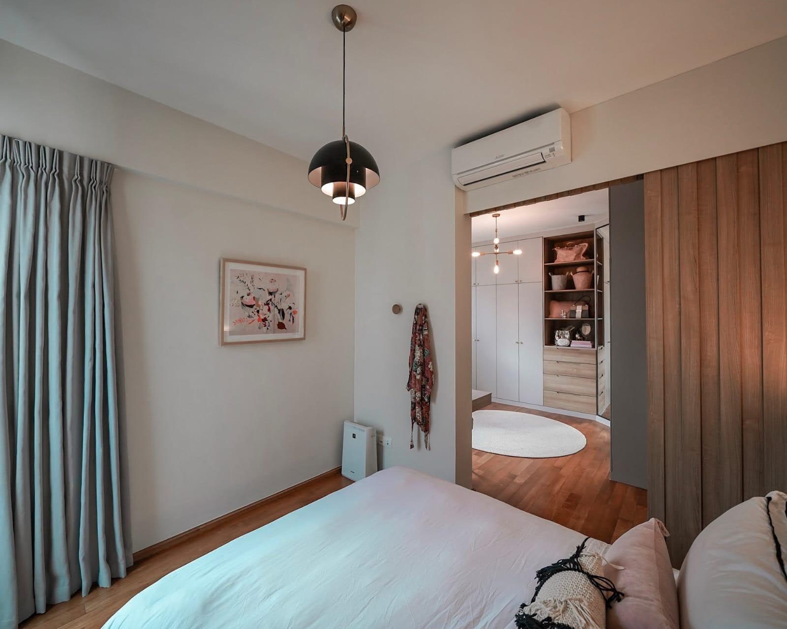 A main bedroom in a Newton condominium with blue-grey curtains, a pendant hung beside the bed, and a fluted timber door standing open onto a lit walk-in wardrobe.