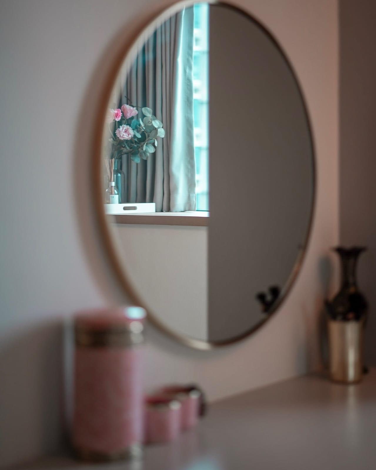 A round mirror above a dressing table in a Newton condominium, reflecting a curtained window with a reed diffuser and stems of pink carnations standing on the sill.