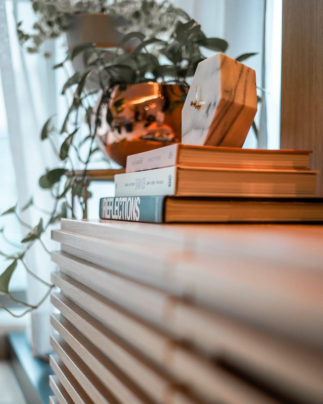 Books stacked on a white slatted console in a Newton condominium, with a hexagonal marble clock resting on them and trailing foliage in a copper planter behind.