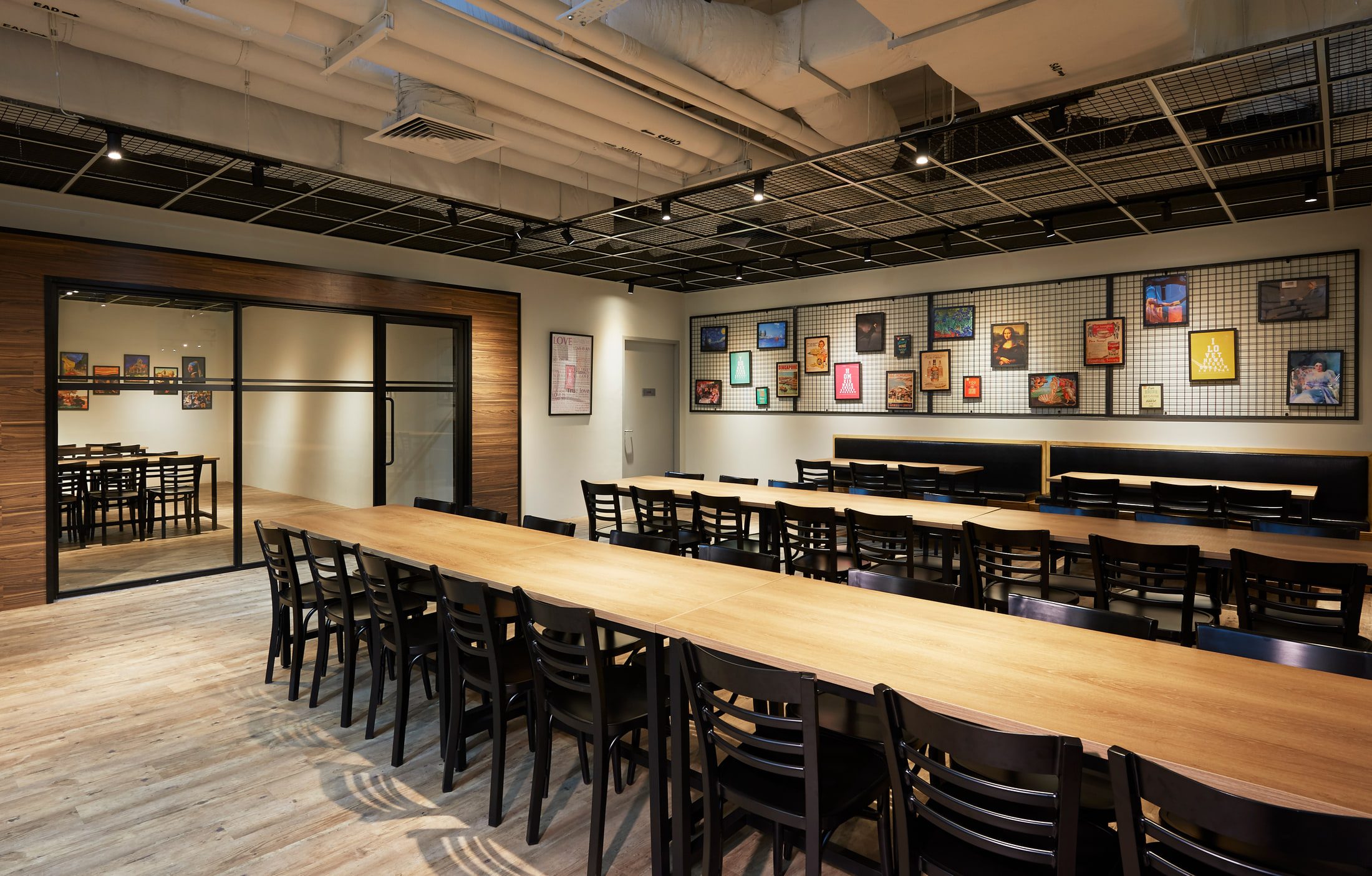 A staff dining hall with long pale timber tables, black bentwood chairs, an exposed services ceiling, and a wall of framed prints hung on black mesh panels.