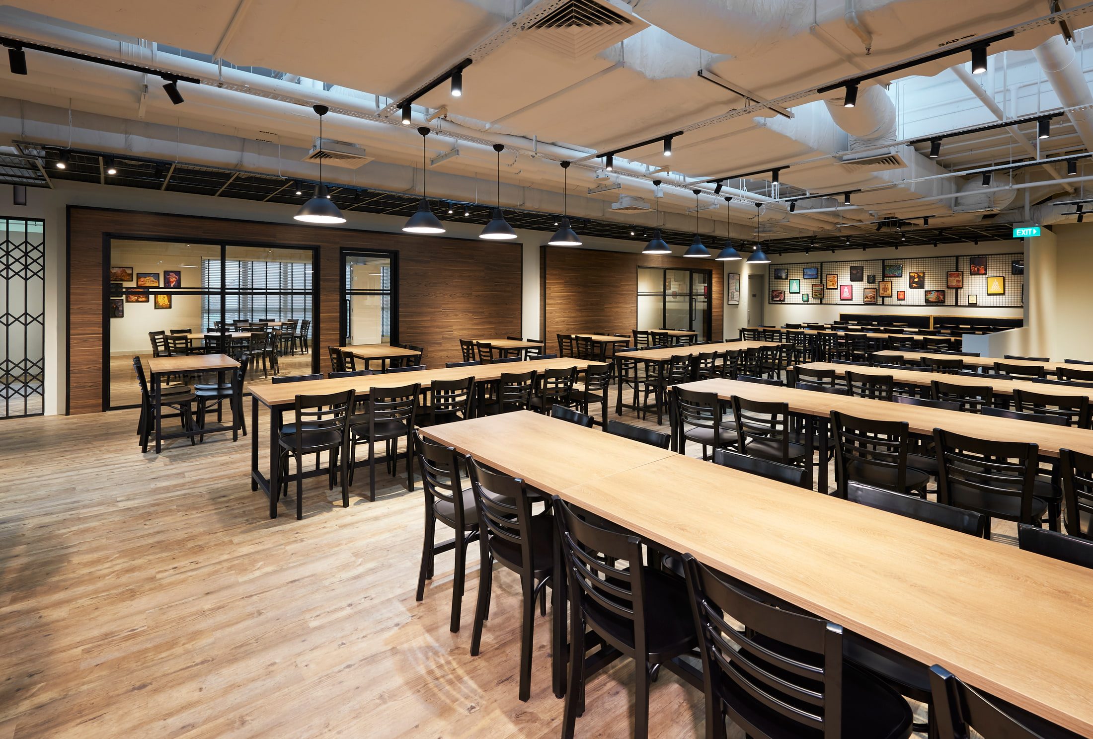 A staff canteen with long pale timber refectory tables and black chairs, a brick wall hung with framed prints, and track lighting under an exposed services ceiling.