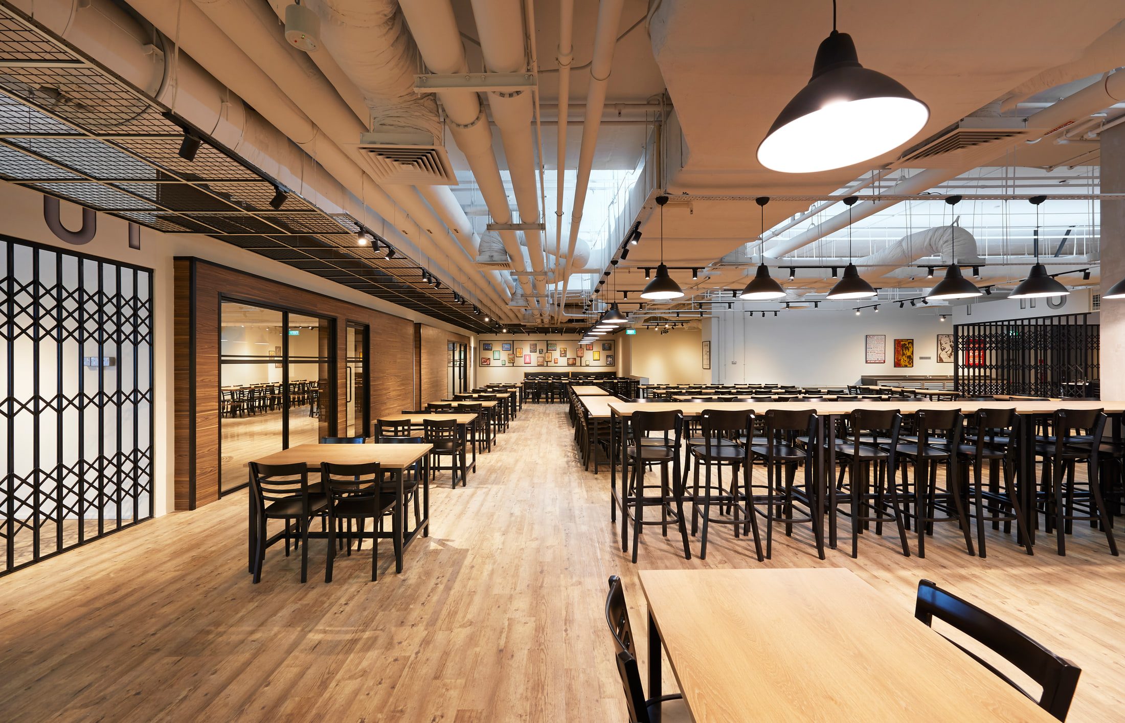 A canteen aisle seen end on, with black dome pendants over communal tables and folding metal grille screens dividing the timber-floored run.