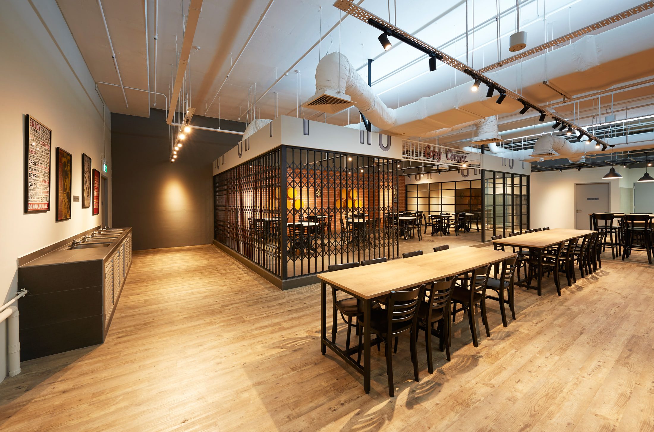 A canteen hall with timber tables and a tiled wash-up counter, screened from a brick-lined games room by a steel scissor gate under exposed ductwork.