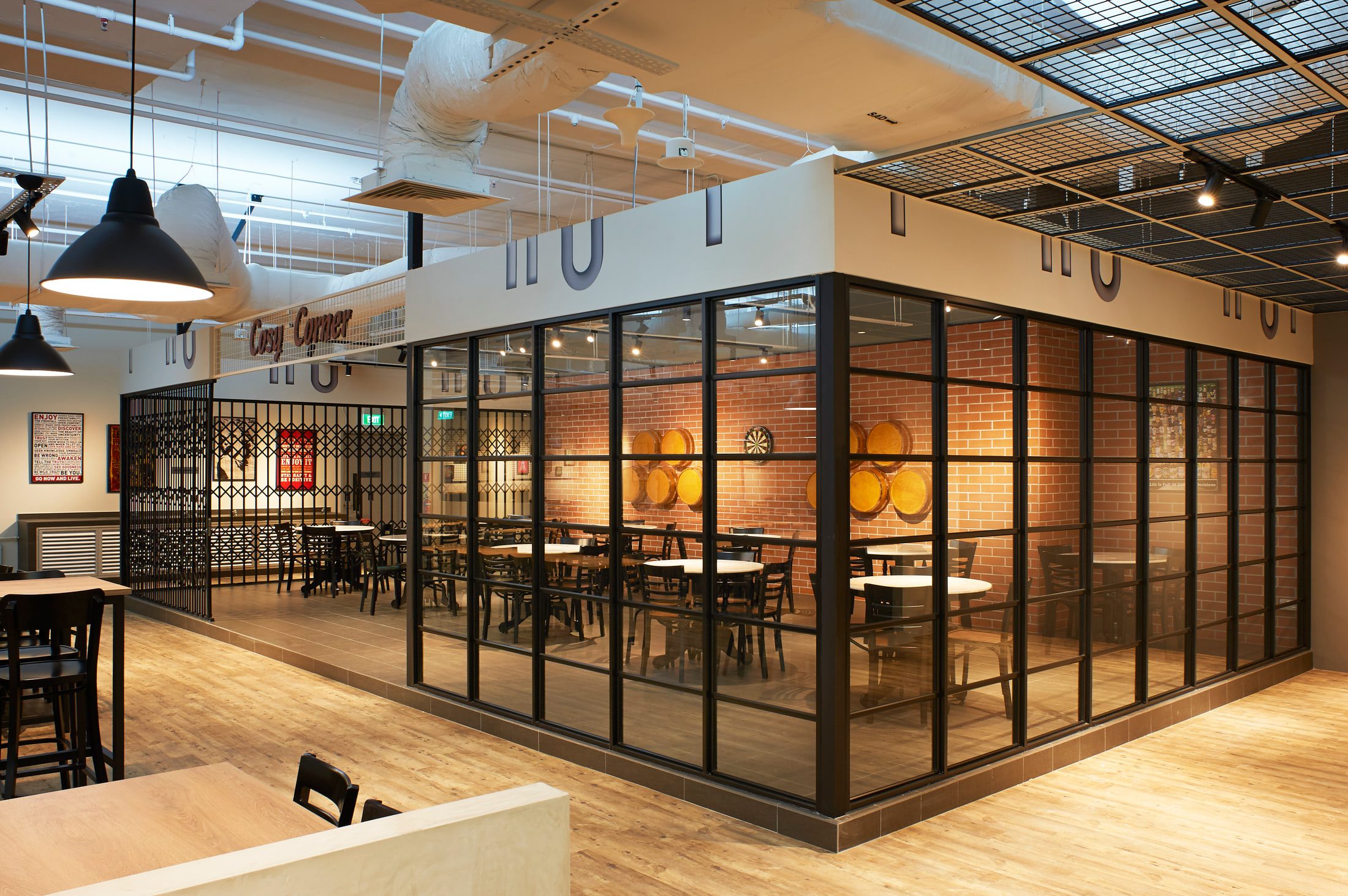 A glazed meeting room framed in black steel against exposed brick, with barrel tables inside and painted lettering across the fascia above.