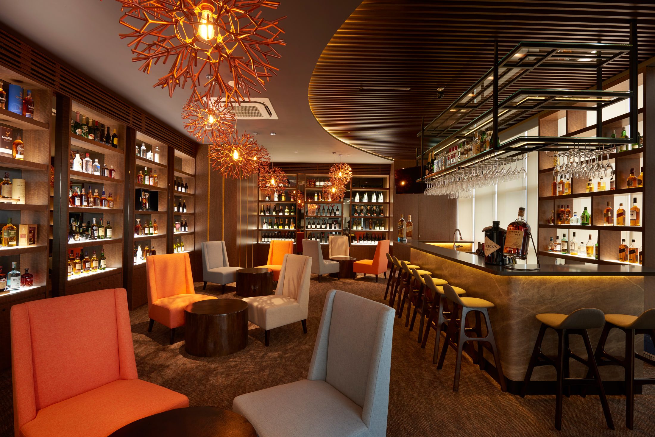 A bar with copper starburst pendants over a stone counter, backlit bottle shelving behind, and orange and cream upholstered chairs on dark carpet.