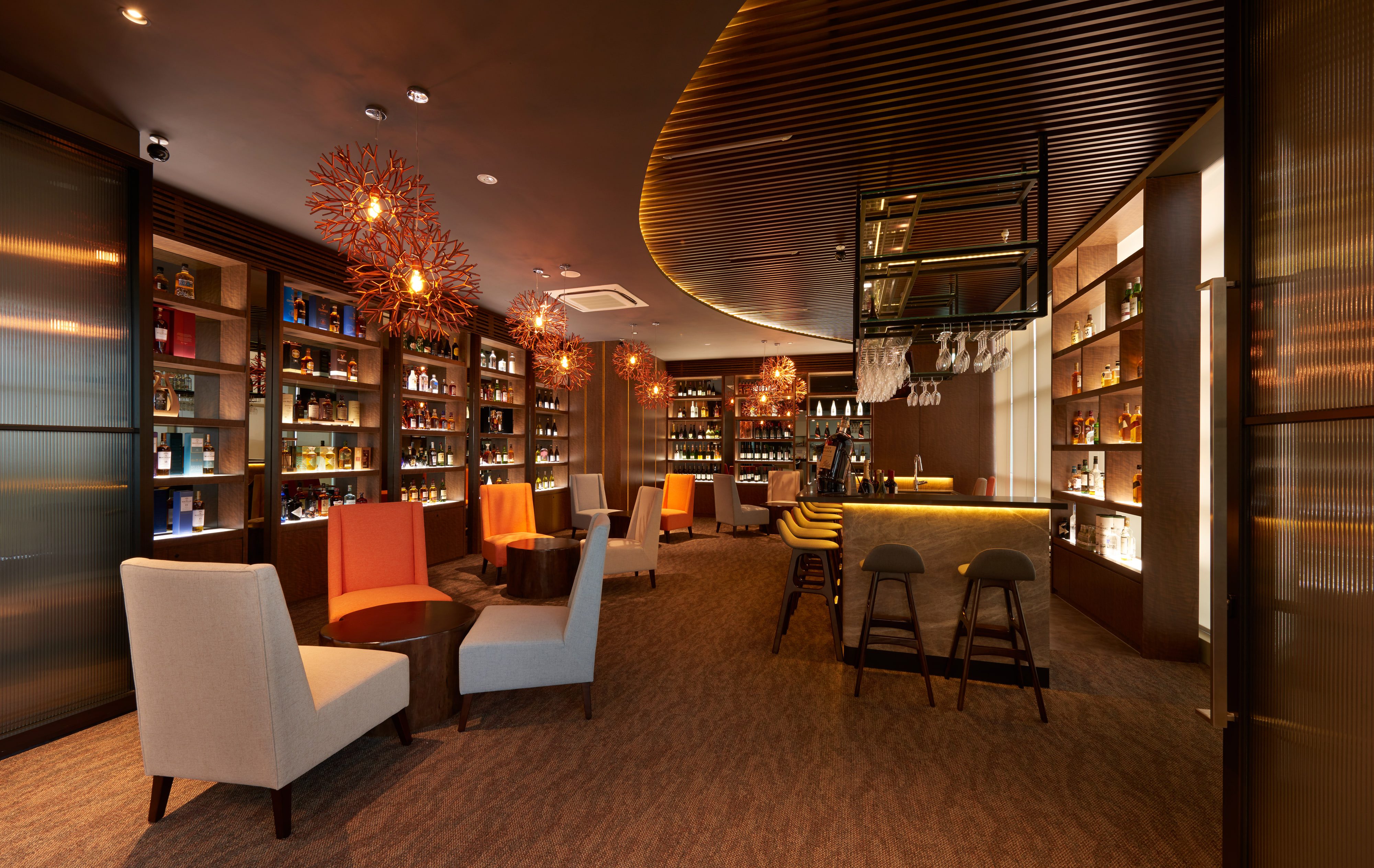 A bar and lounge with a backlit stone counter, timber bottle shelving, branching pendant lamps and coral and oatmeal armchairs on textured carpet.