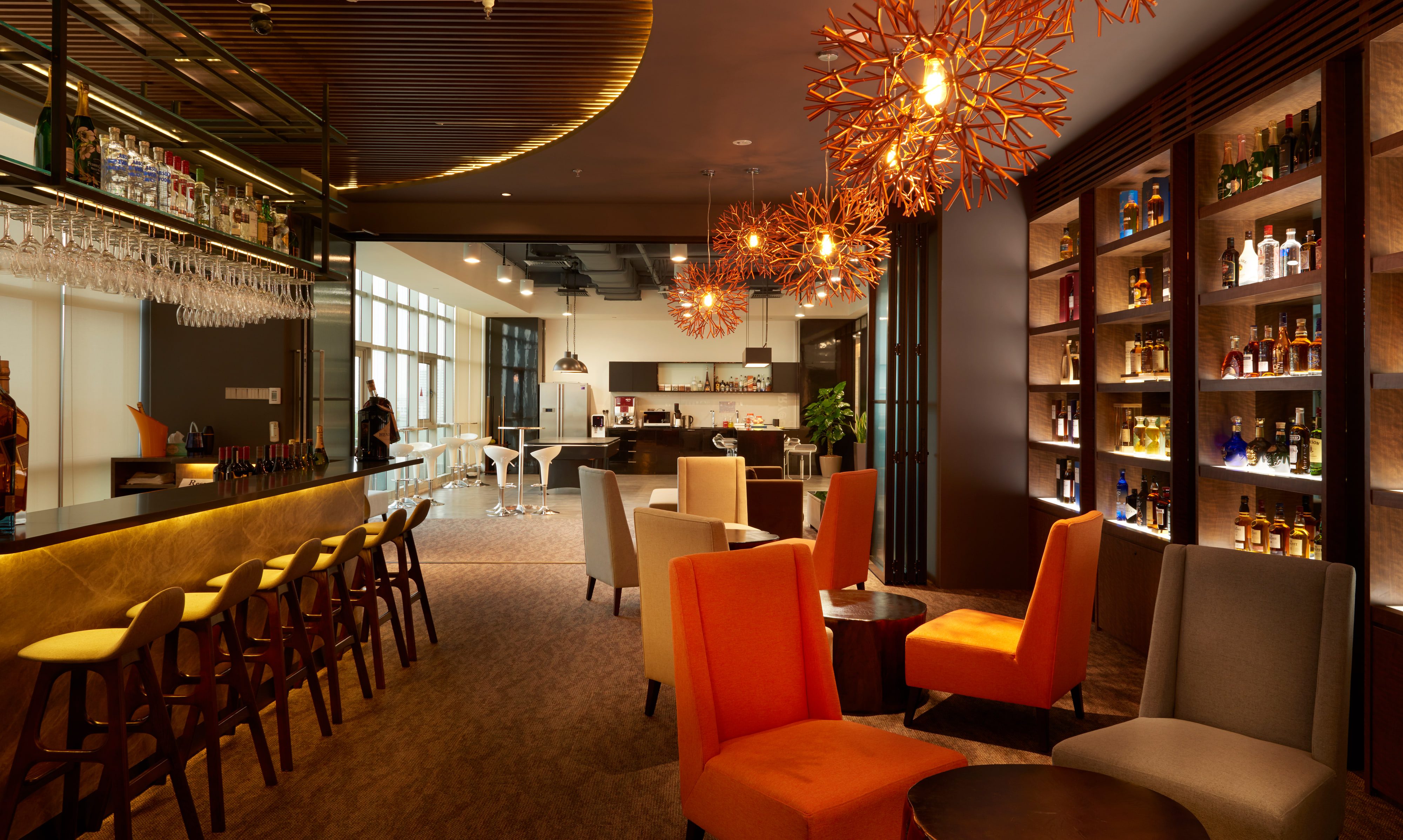 A bar counter with mirrored back shelving and mustard-topped stools, opening past a folding glazed partition to a daylit pantry with a poseur table.