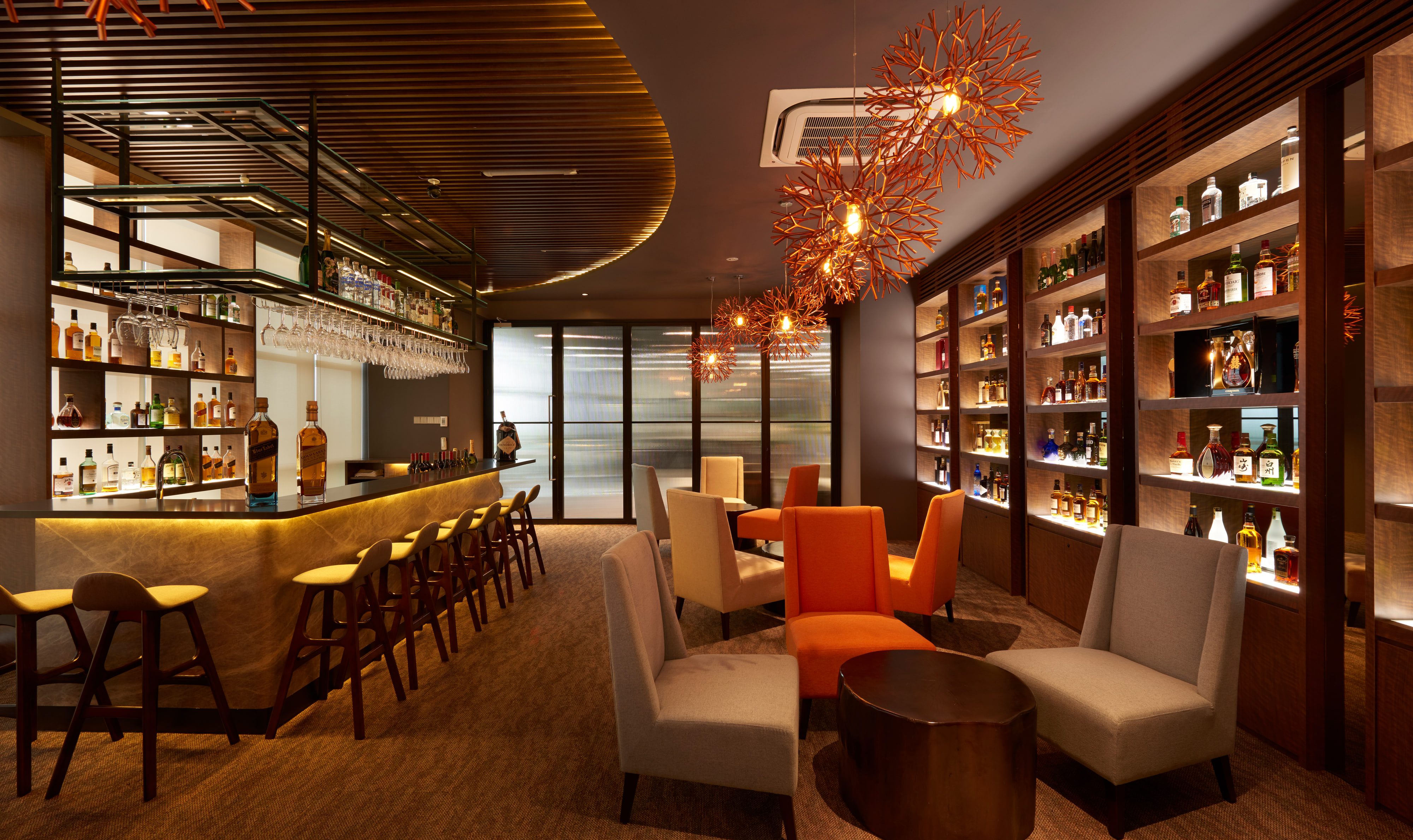 An angled bar counter in backlit veined stone with a black top, timber stools along its face, and lit shelving running the full height of the wall opposite.