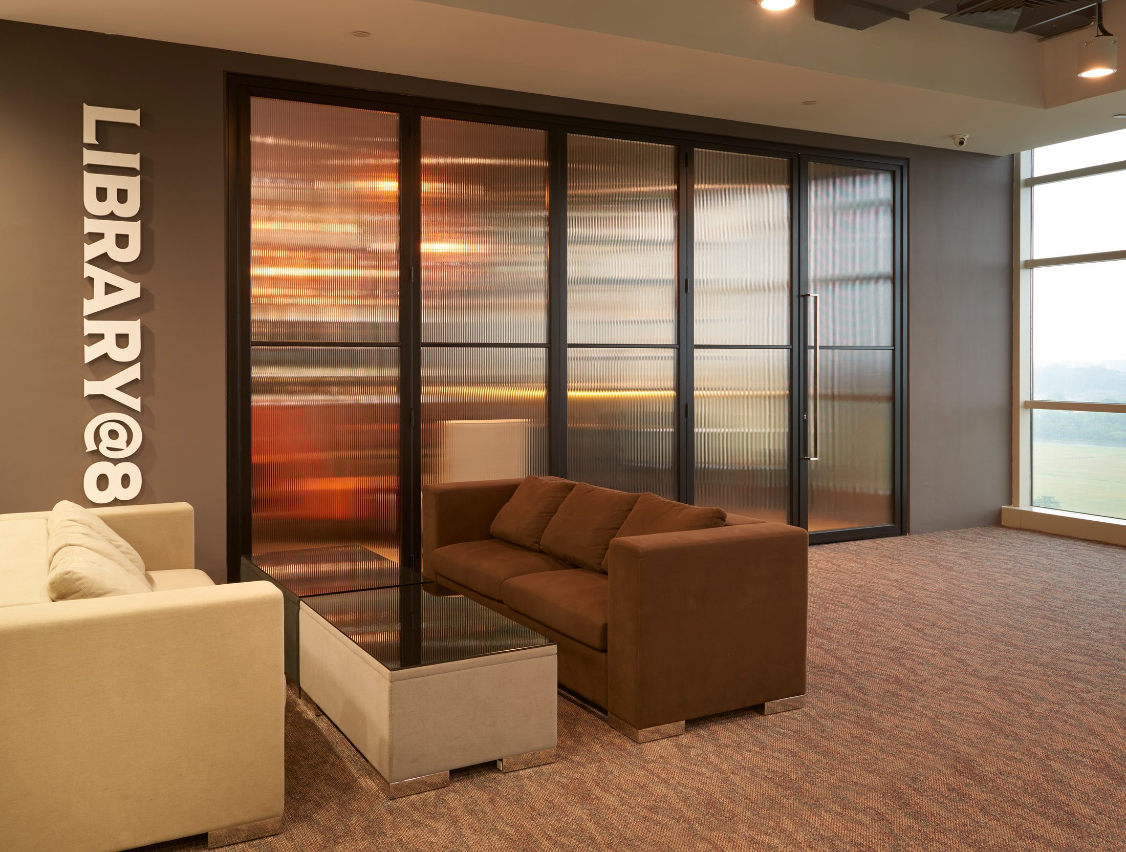 A glazed lounge signed as a library, with brown and cream sofas set around a concrete planter behind reeded-glass sliding doors.