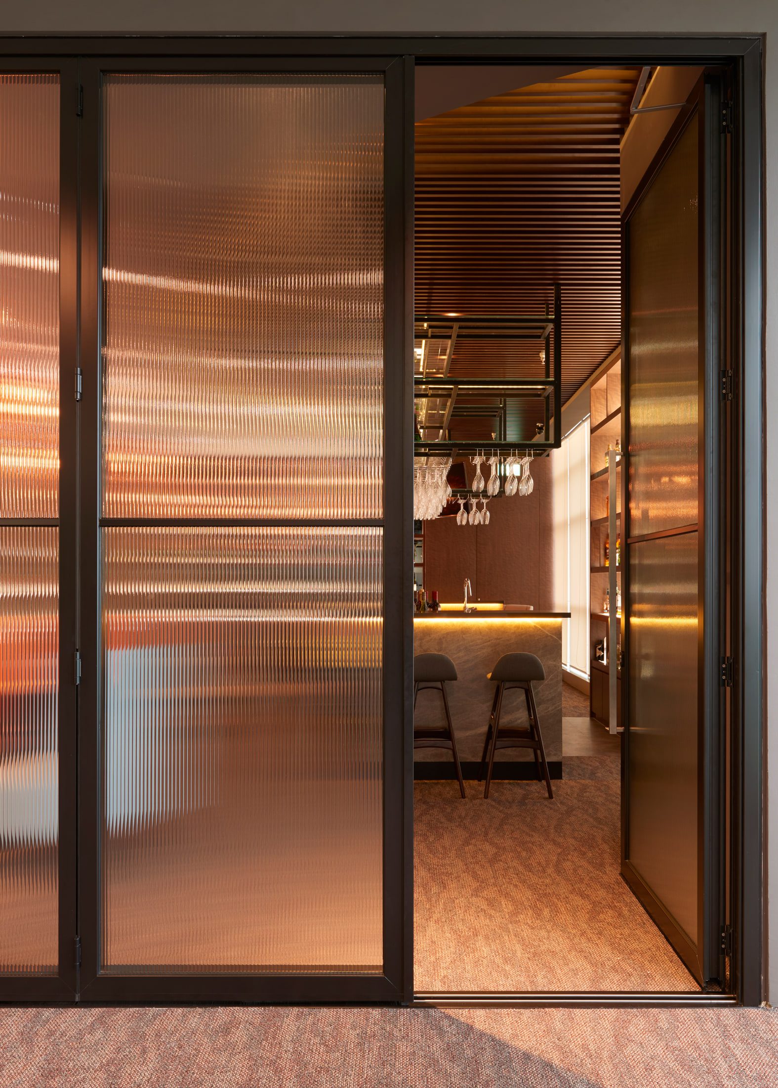 A reeded-glass and dark metal partition door opening into the bar, with stools and hanging glassware visible through it.