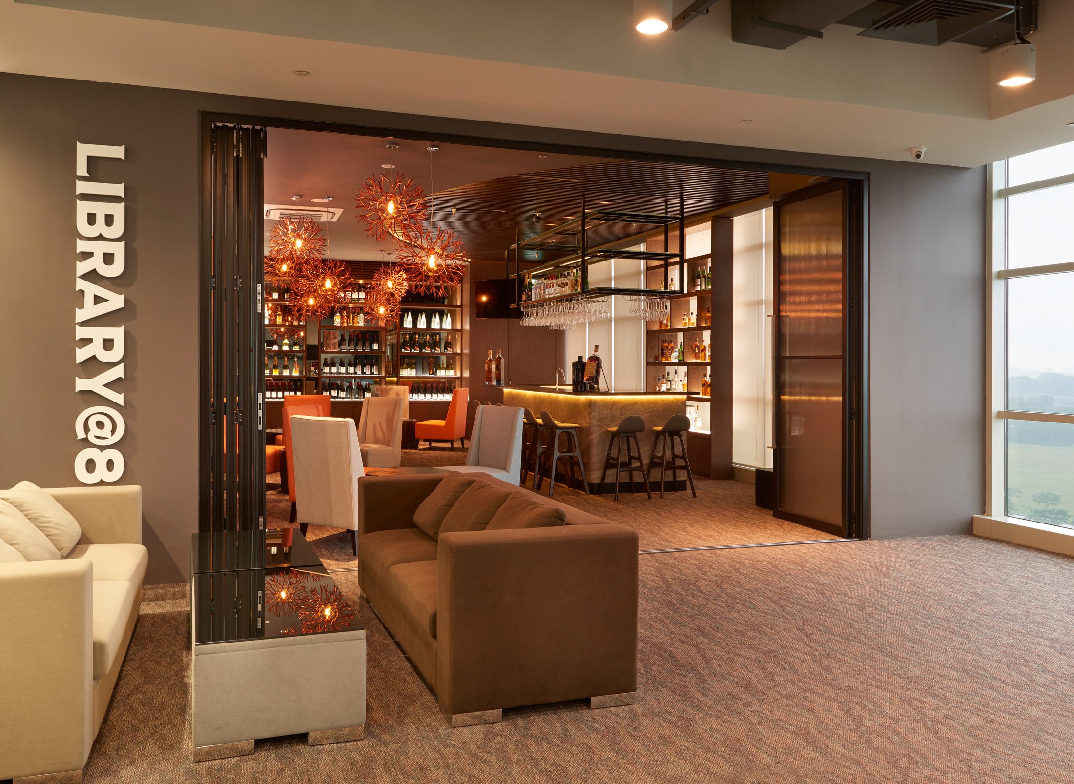 A lounge with cream and taupe sofas on patterned carpet beside a full-height window, looking through folding bronze-glazed doors into a warmly lit bar.