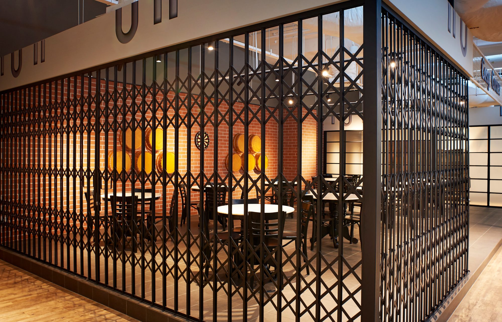 A folding diamond-pattern metal grille drawn across a brick-walled dining bay, tables and barrels showing through the mesh.