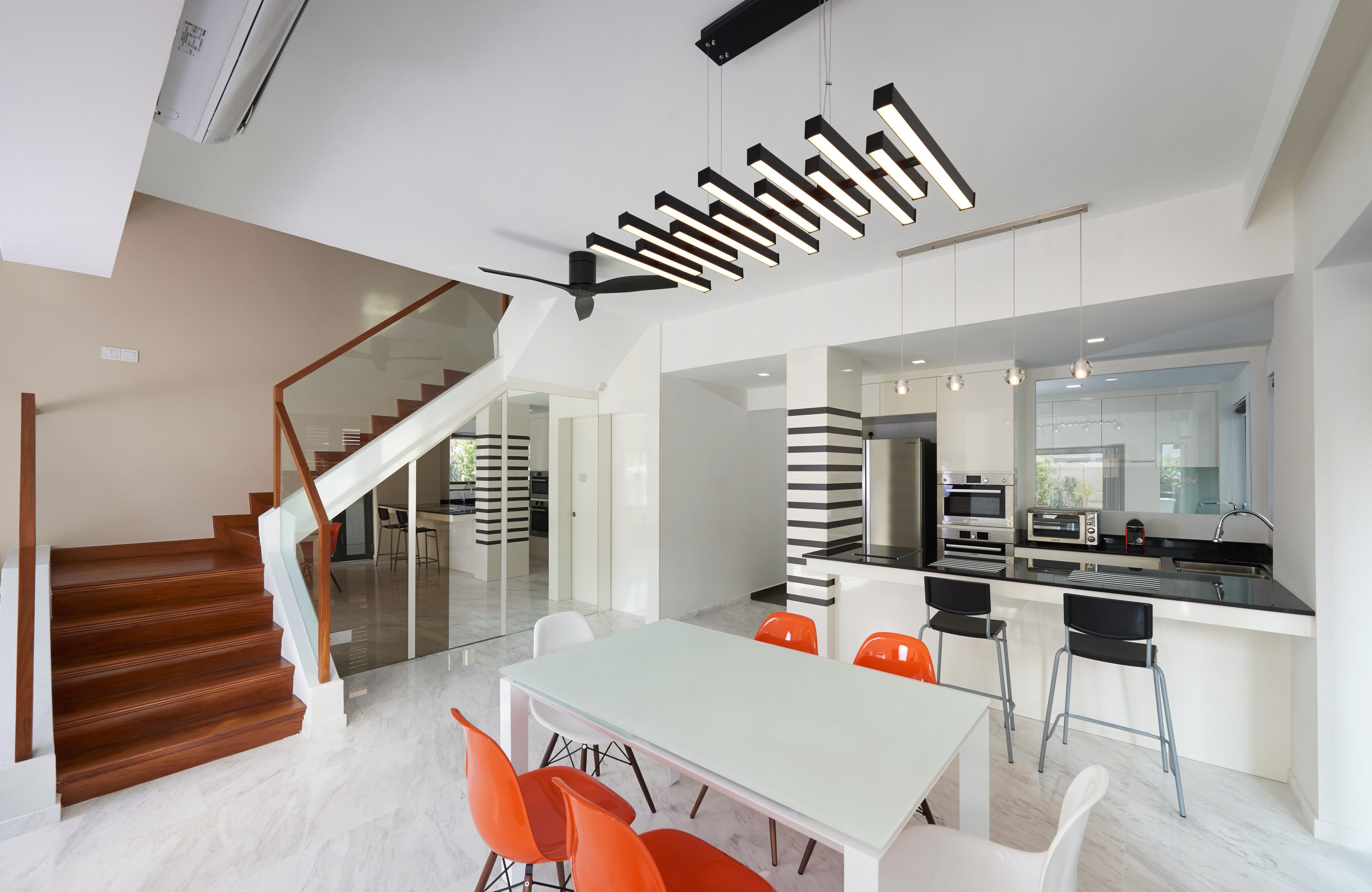 An open dining and kitchen level with a glass-topped table, orange moulded chairs, a black-and-white striped column and a varnished timber stair rising behind a glass balustrade.