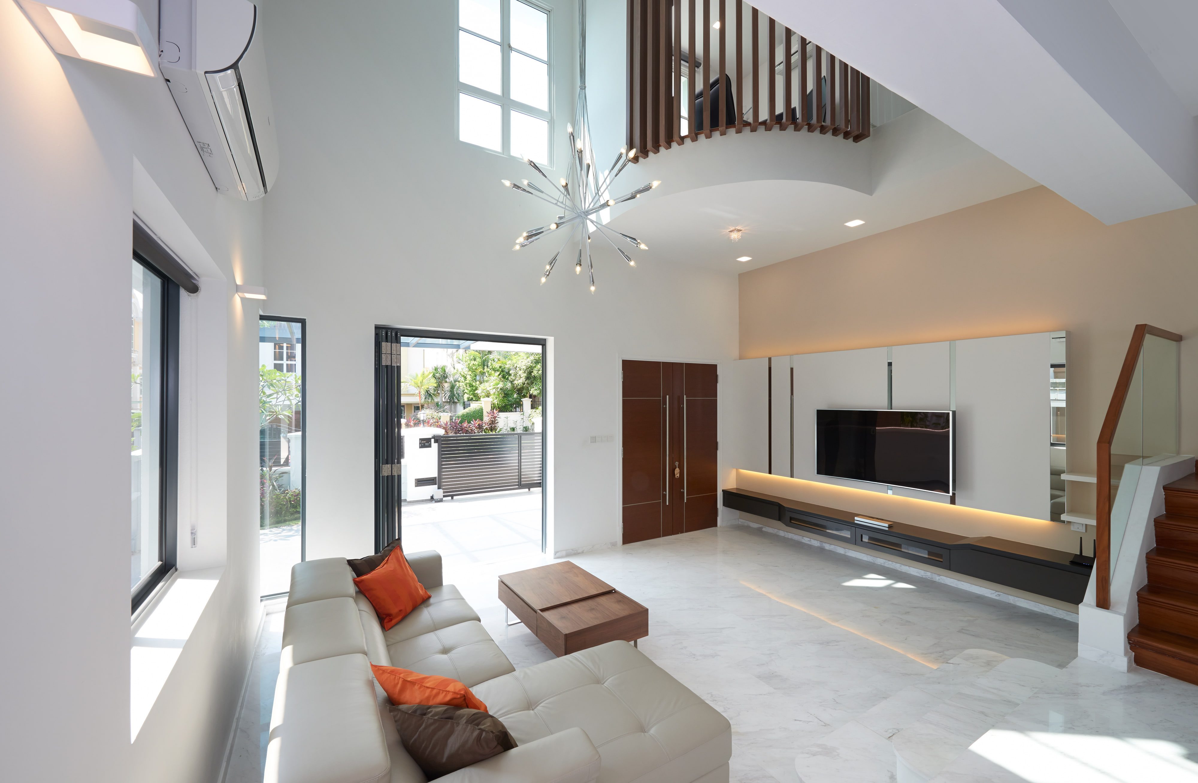 A double-height living room with a cream leather sofa, a full-width media wall lit from behind, marble flooring and folding glazed doors open to a covered car porch.