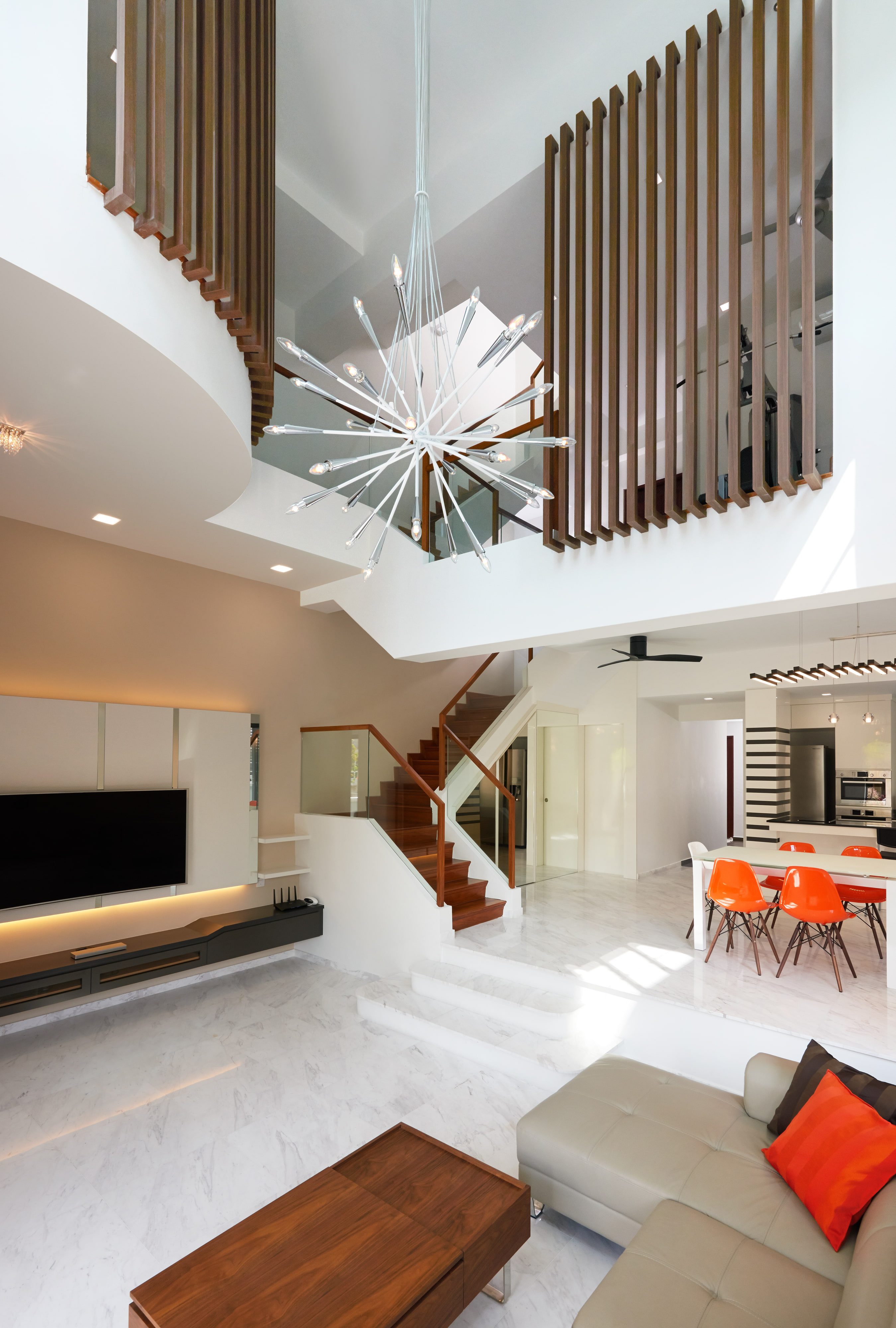 A double-height living space with a starburst pendant hung in the void, timber slat screens to the upper landing, and a dining table with orange chairs beyond.