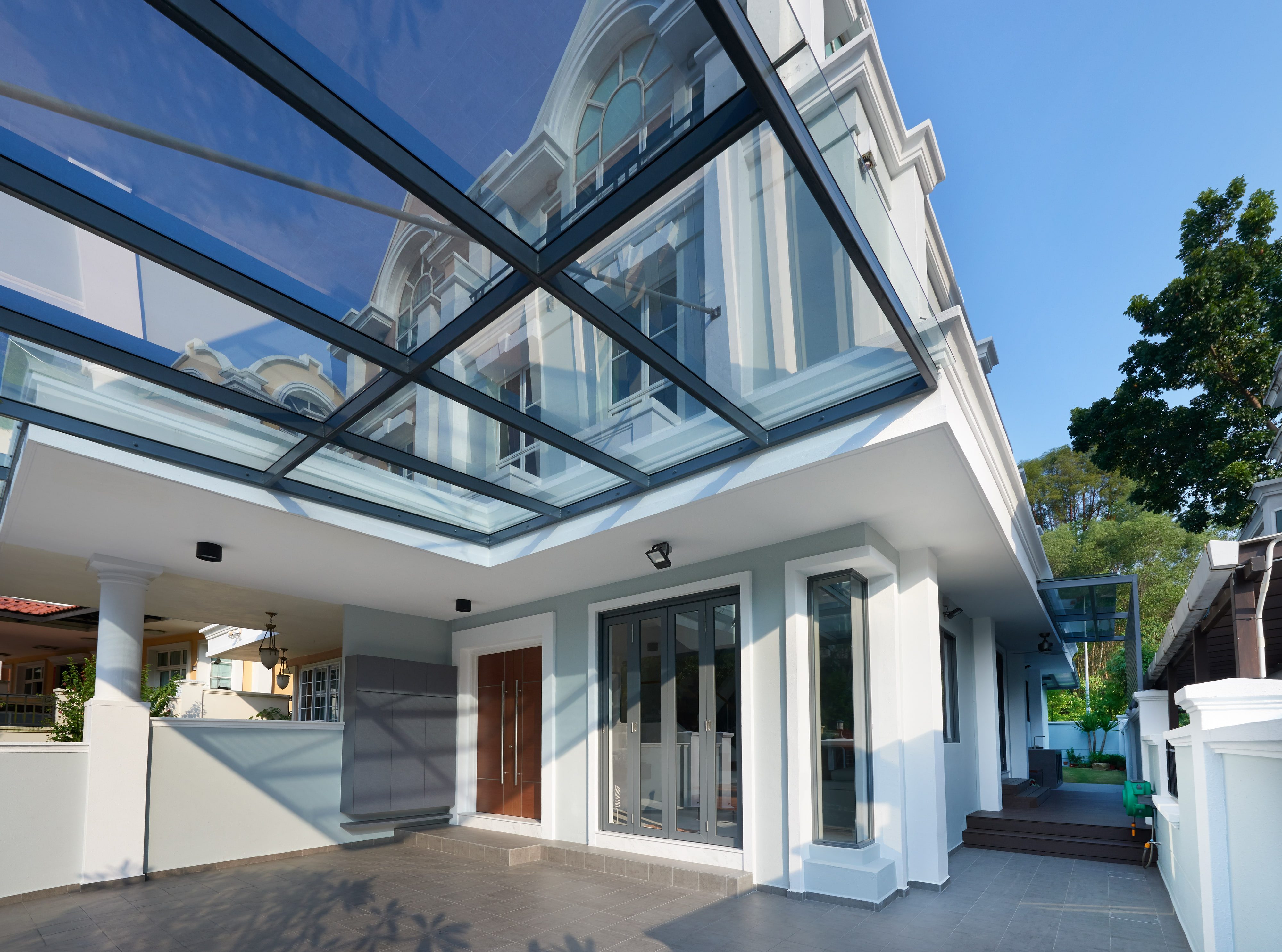 A car porch under a flat steel-framed glass canopy, with a timber entrance door, grey aluminium folding doors and the original moulded gable rising above.