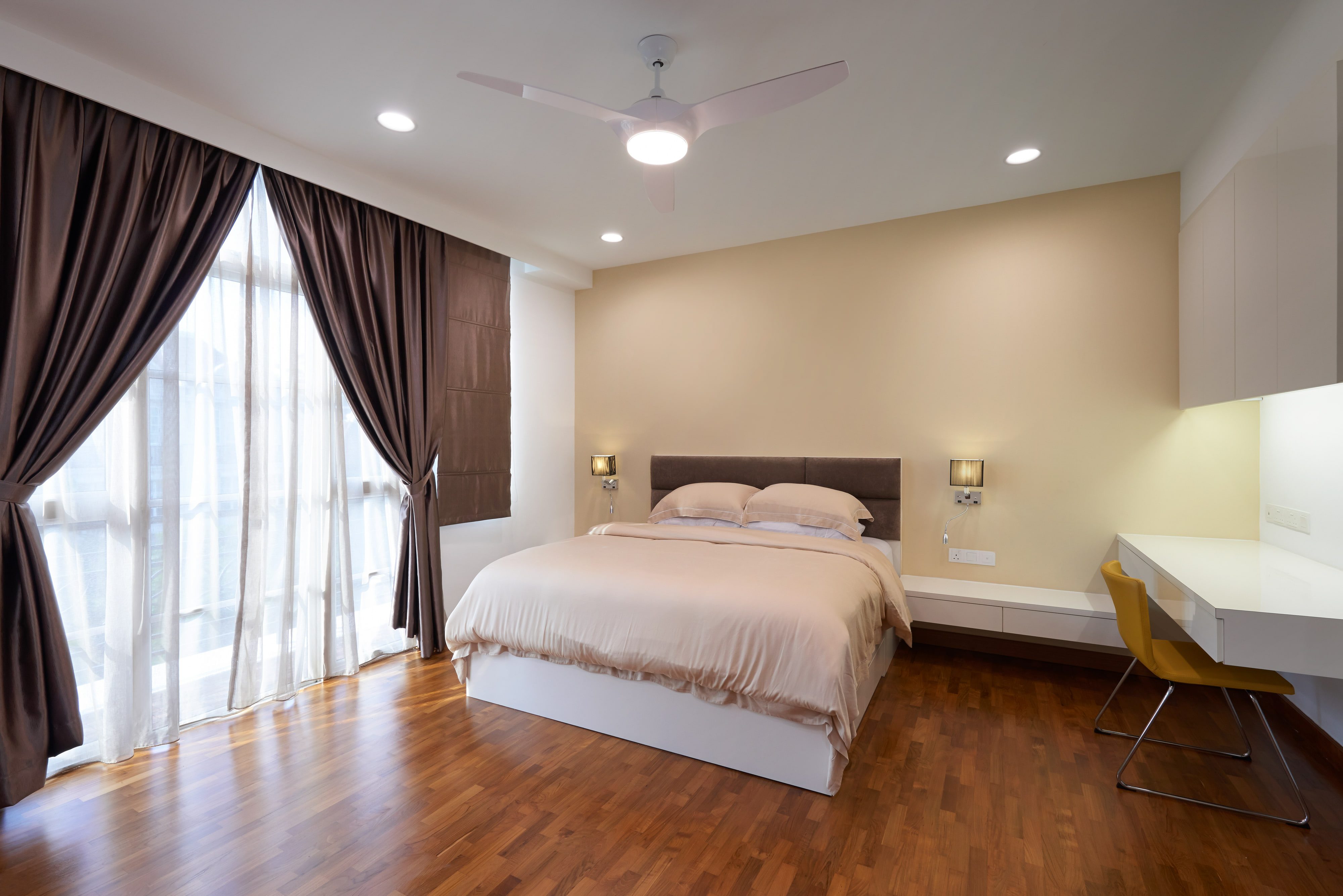 A bedroom with a grey upholstered headboard, brown silk curtains over a full-height window, dark timber strip flooring and a white lacquer desk with a mustard chair.