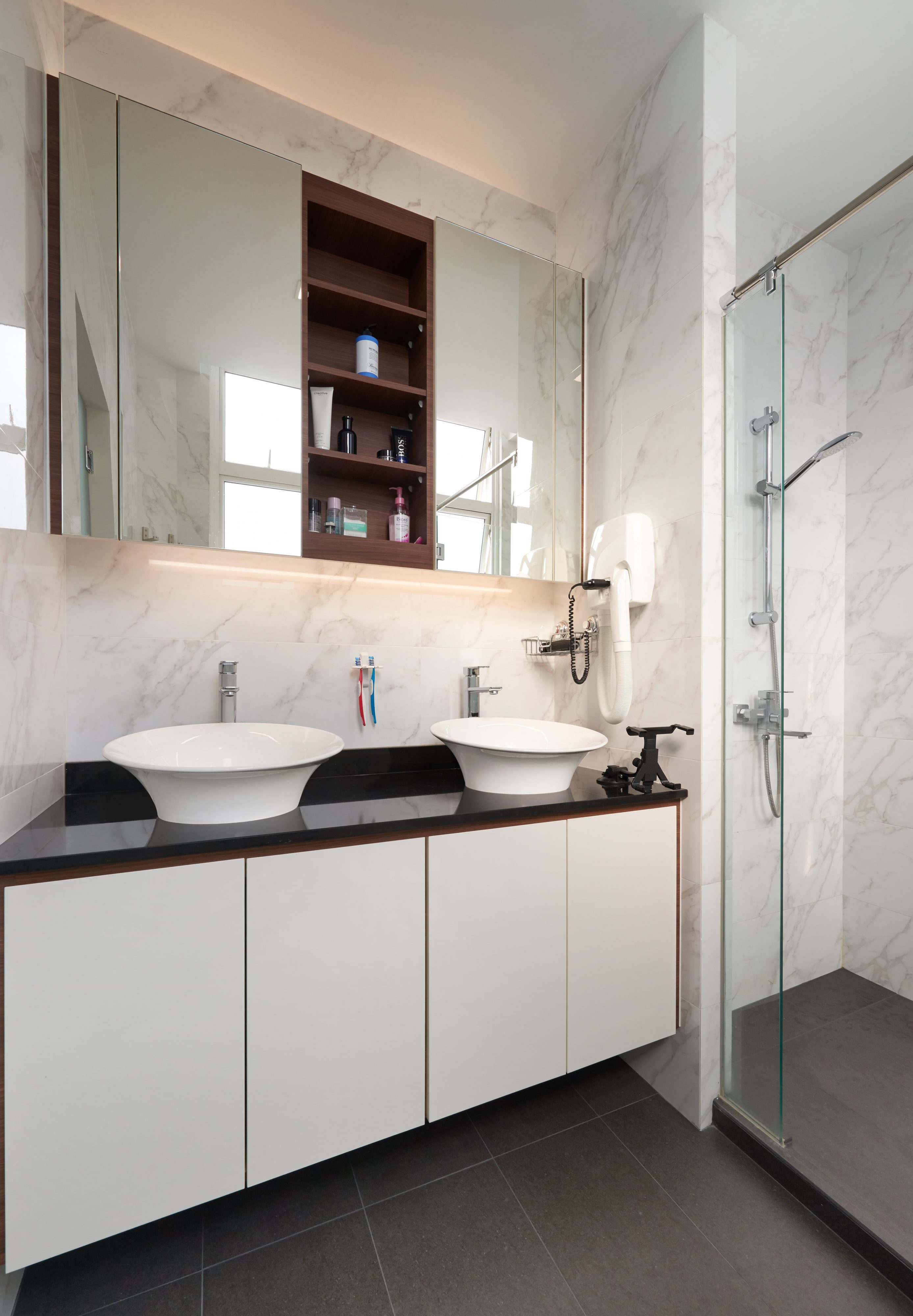 A bathroom with twin white vessel basins on a black stone top over white lacquer drawers, a walnut open shelf set between mirrored cabinets and a glazed shower alongside.