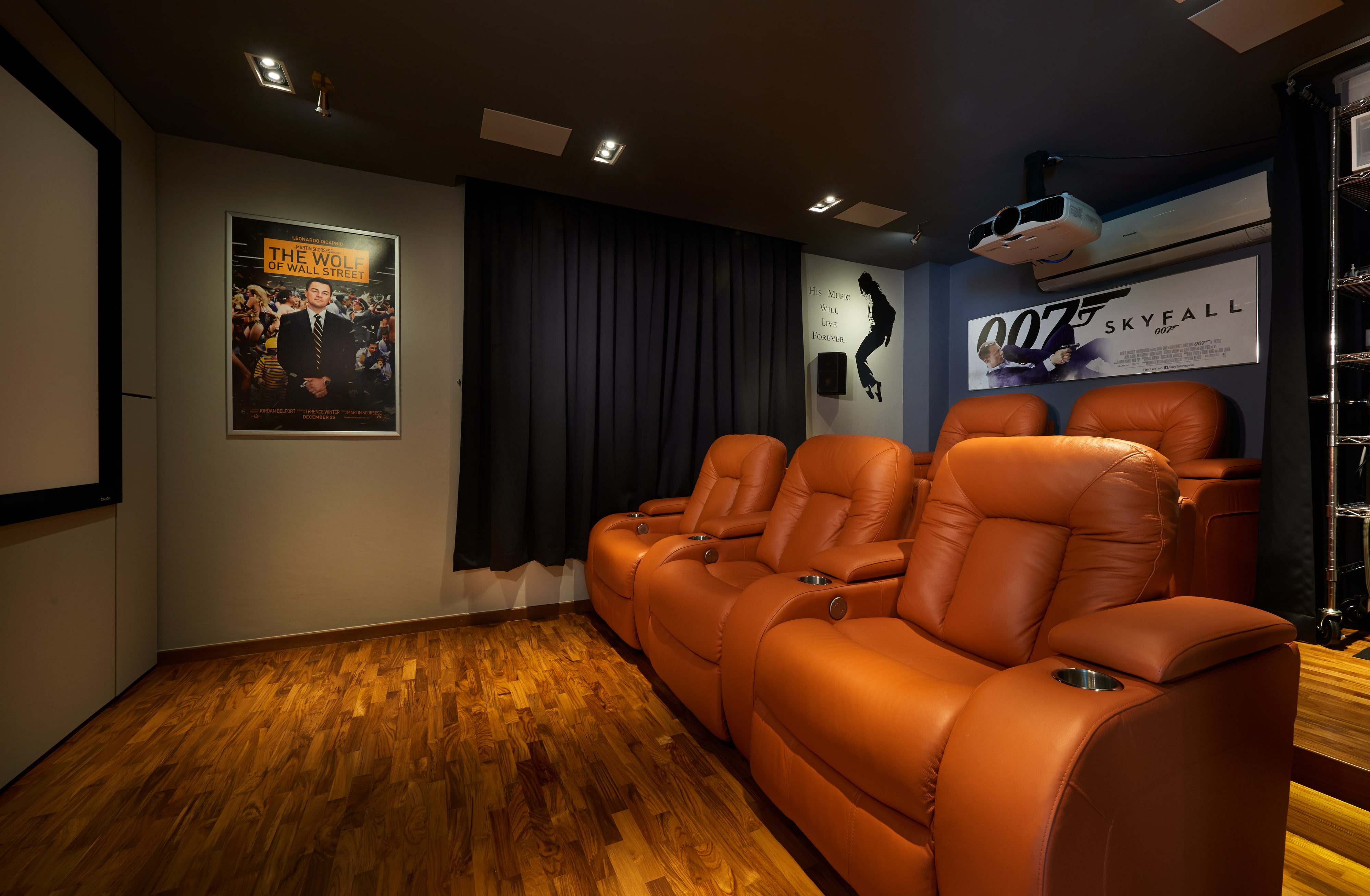 A home cinema with two rows of tan leather recliners facing a projection screen, blackout curtaining, framed film posters and a ceiling-mounted projector.