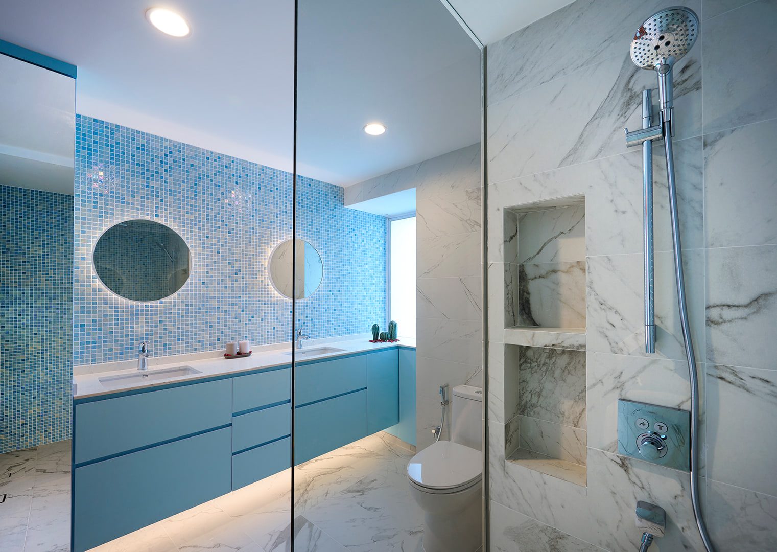 A bathroom with a blue mosaic wall behind twin round mirrors, a pale blue vanity, and a marble-lined shower beyond a glass screen.