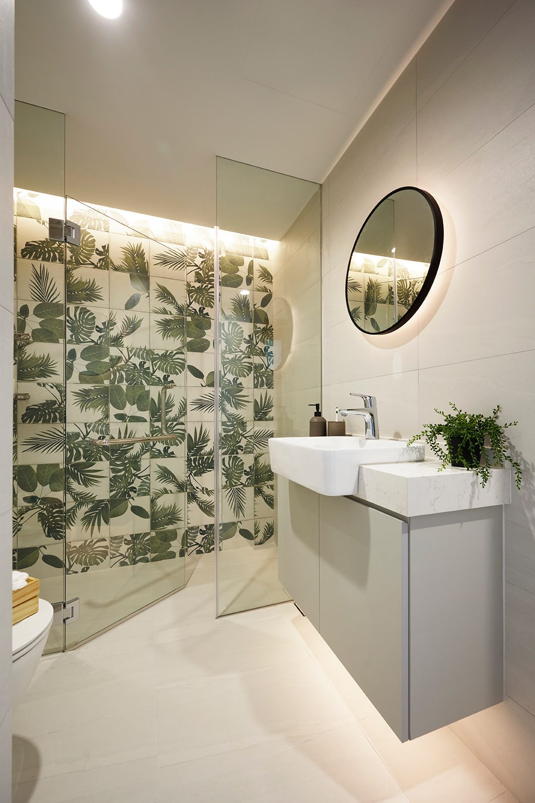 A bathroom with a botanical leaf-patterned tile panel in the shower, a round black-framed mirror, and a stone basin on a pale vanity.