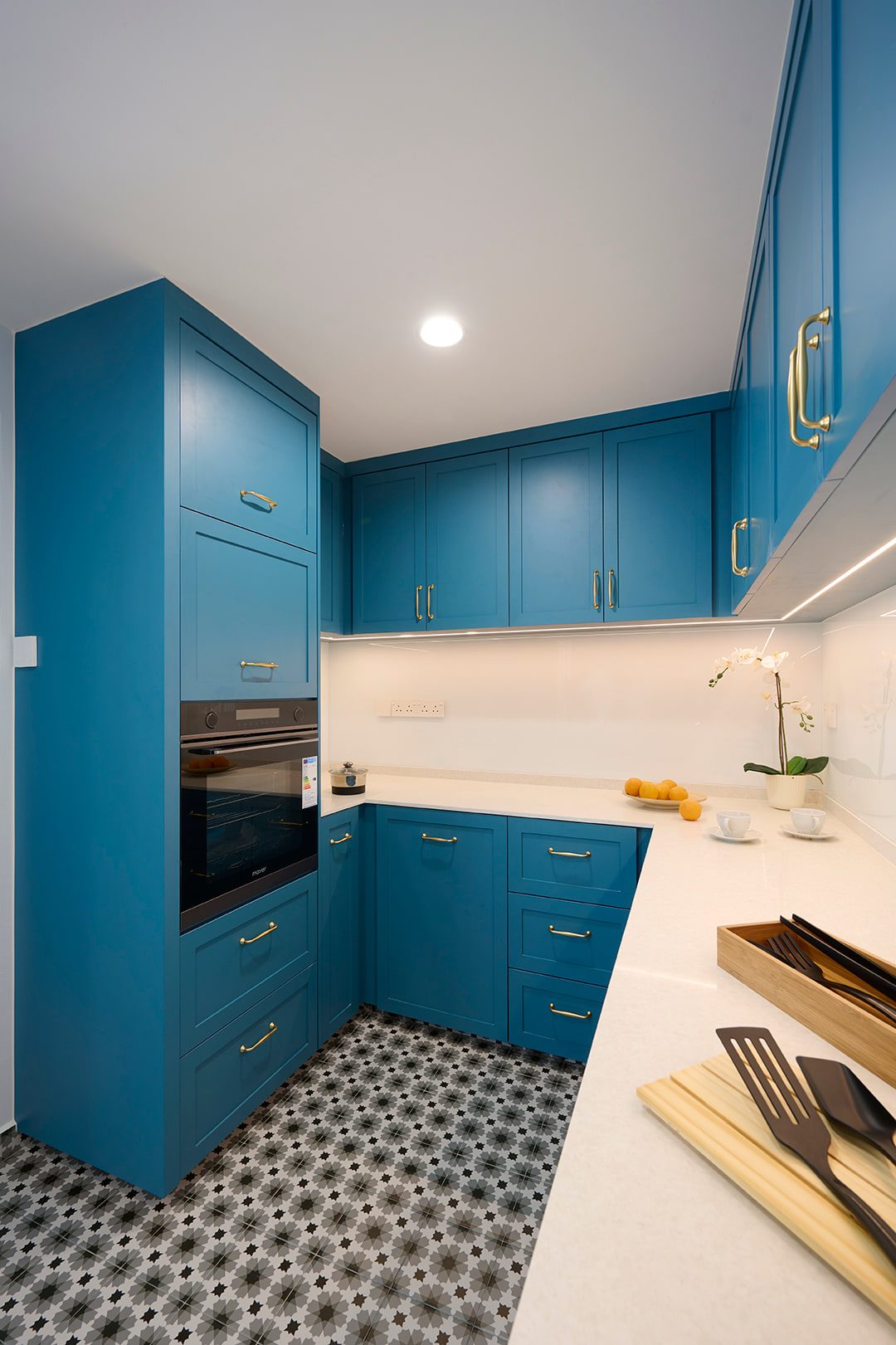 A galley kitchen in blue shaker cabinetry with brass handles, a white worktop and a black and white patterned floor tile.