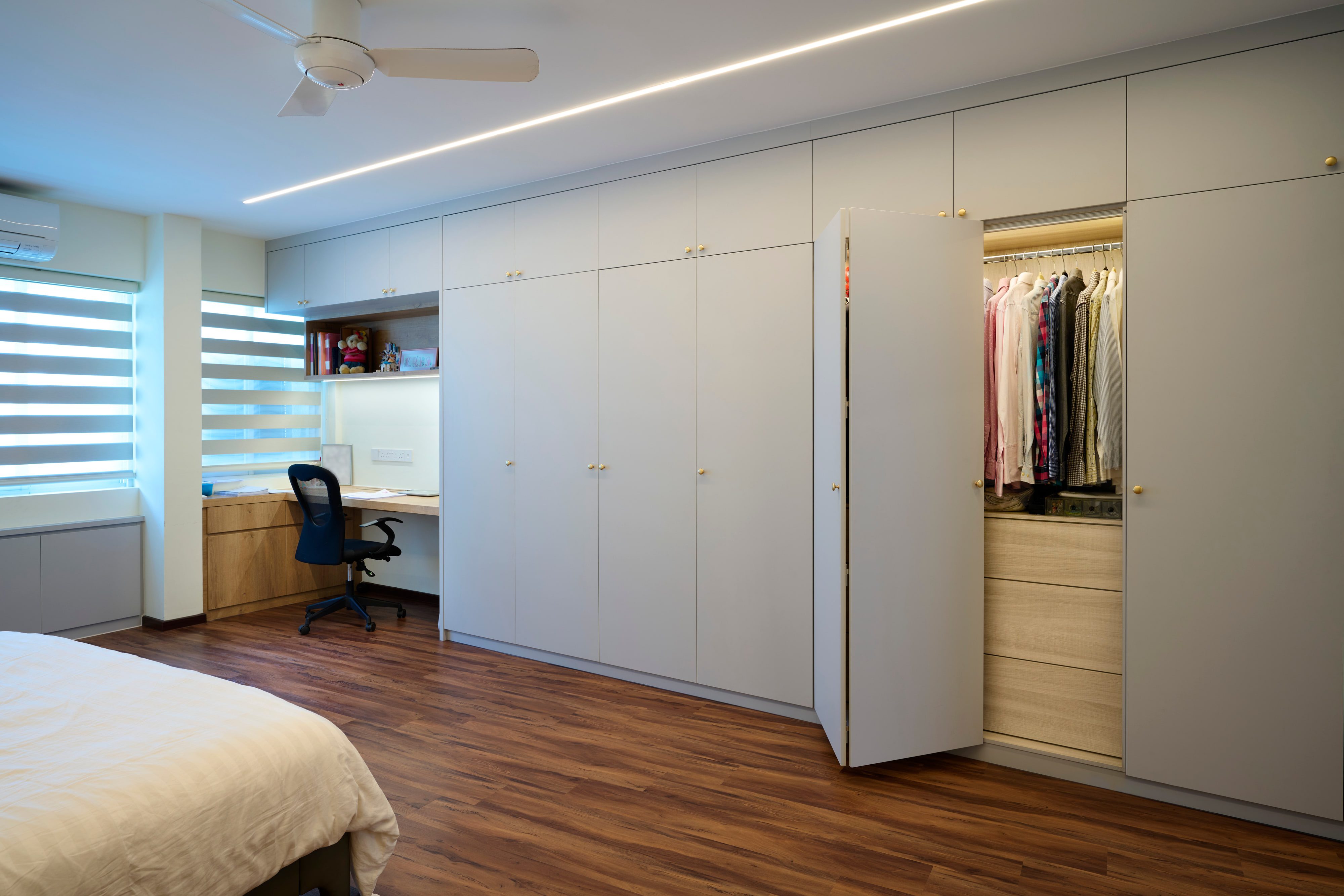 A floor-to-ceiling greige wardrobe wall on brass knobs, one door open to a timber-lined interior, beside a recessed desk under day-night blinds.