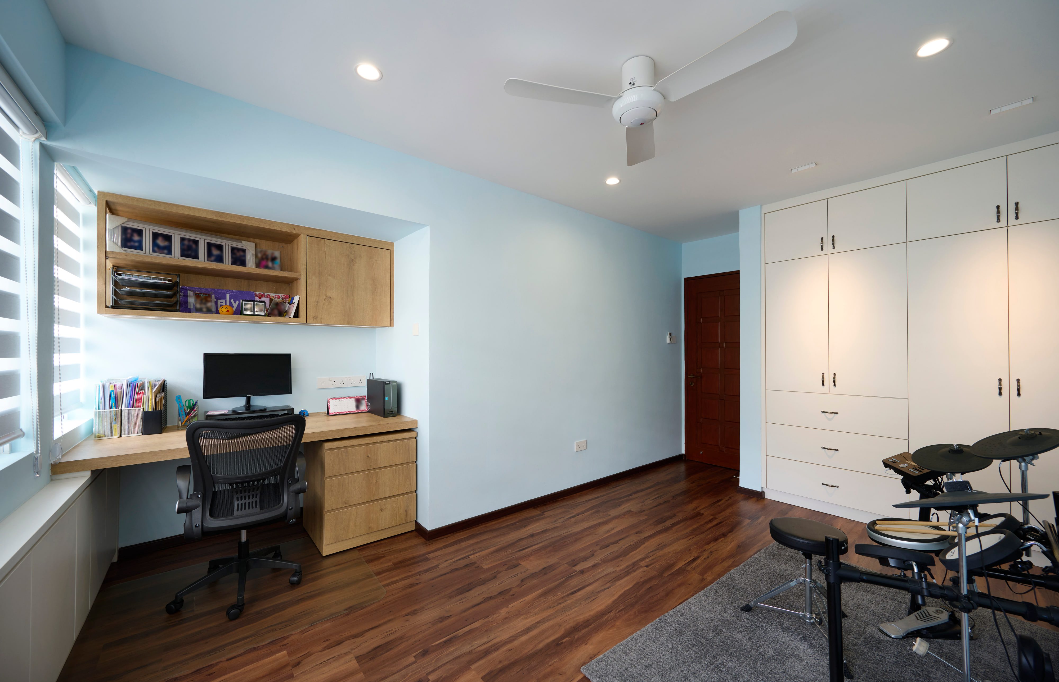 A study in pale blue with a timber desk running from the window sill, a wall-hung cabinet, a white wardrobe with bronze handles and a drum kit.
