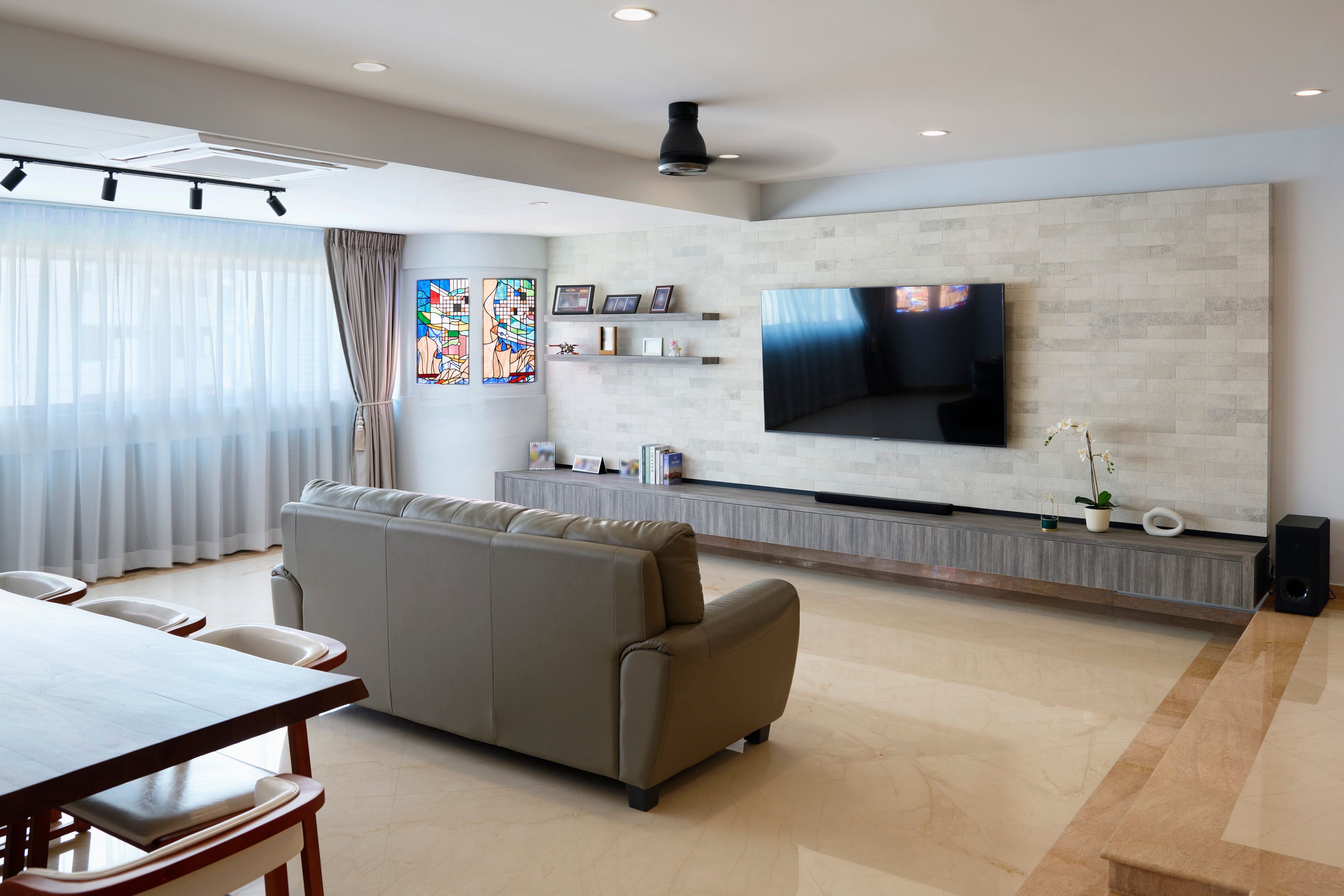A living room with a pale stacked-stone television wall, a long floating console, a taupe leather sofa and stained-glass panels set into a curved wall.