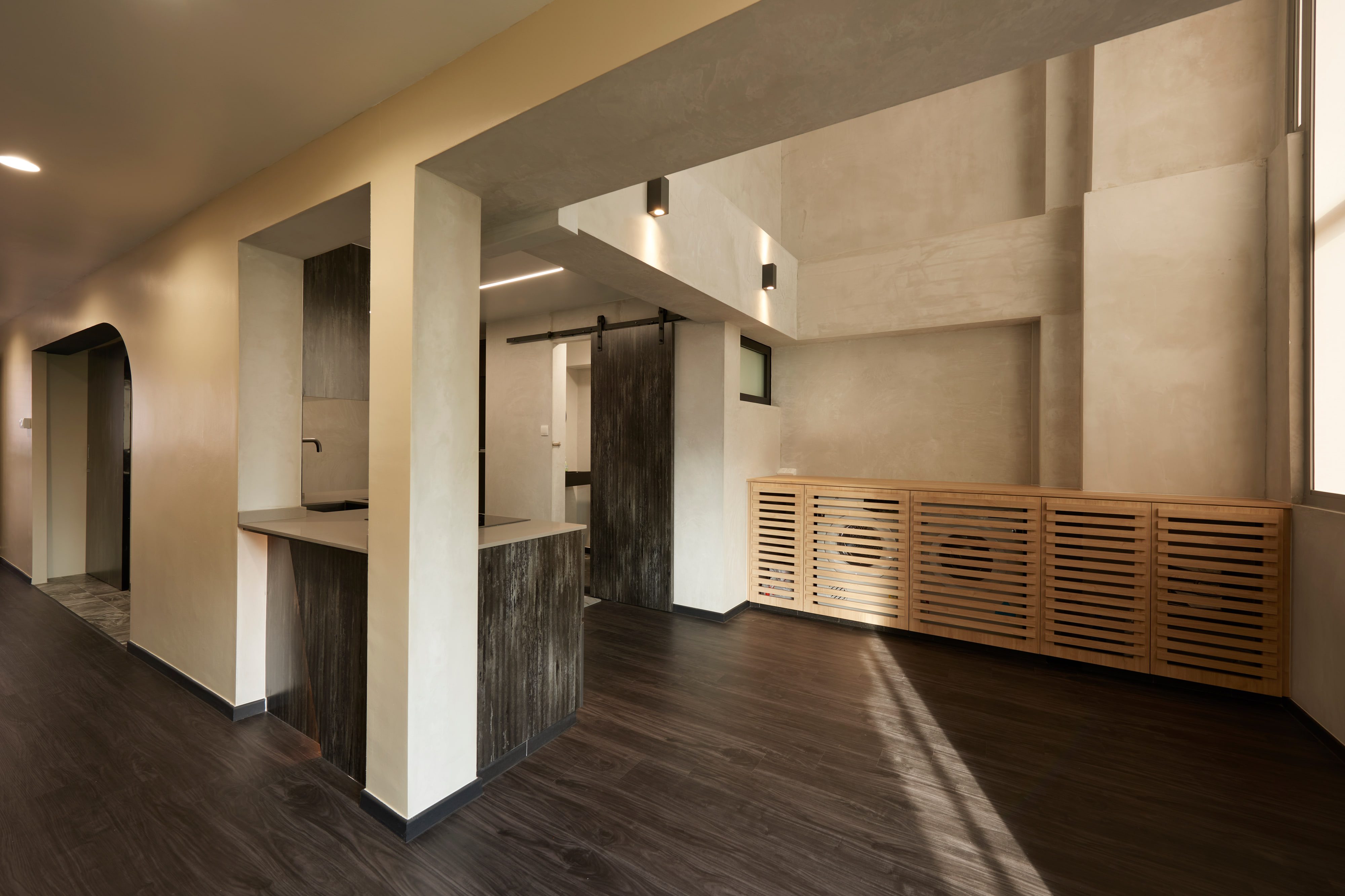 A troweled cement-plaster living room with a dark plank floor, a stone-topped cooking peninsula clad in scorched timber, and a low run of slatted bamboo cabinets under a shaft of afternoon light.