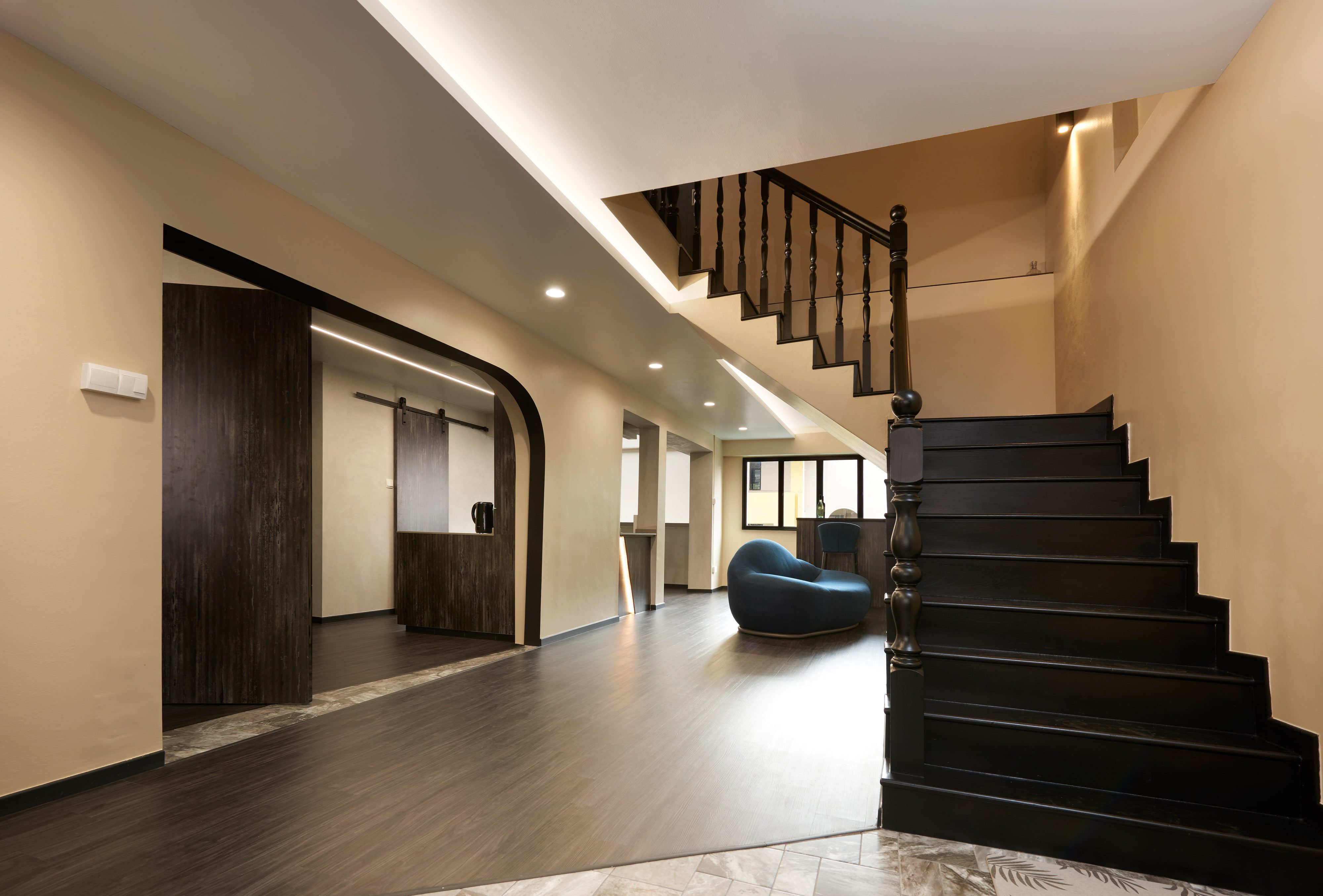 A hall lit from a recessed ceiling cove, the arched opening to the kitchen half closed by a sliding panel in dark scorched timber, with a black turned-baluster stair at right.