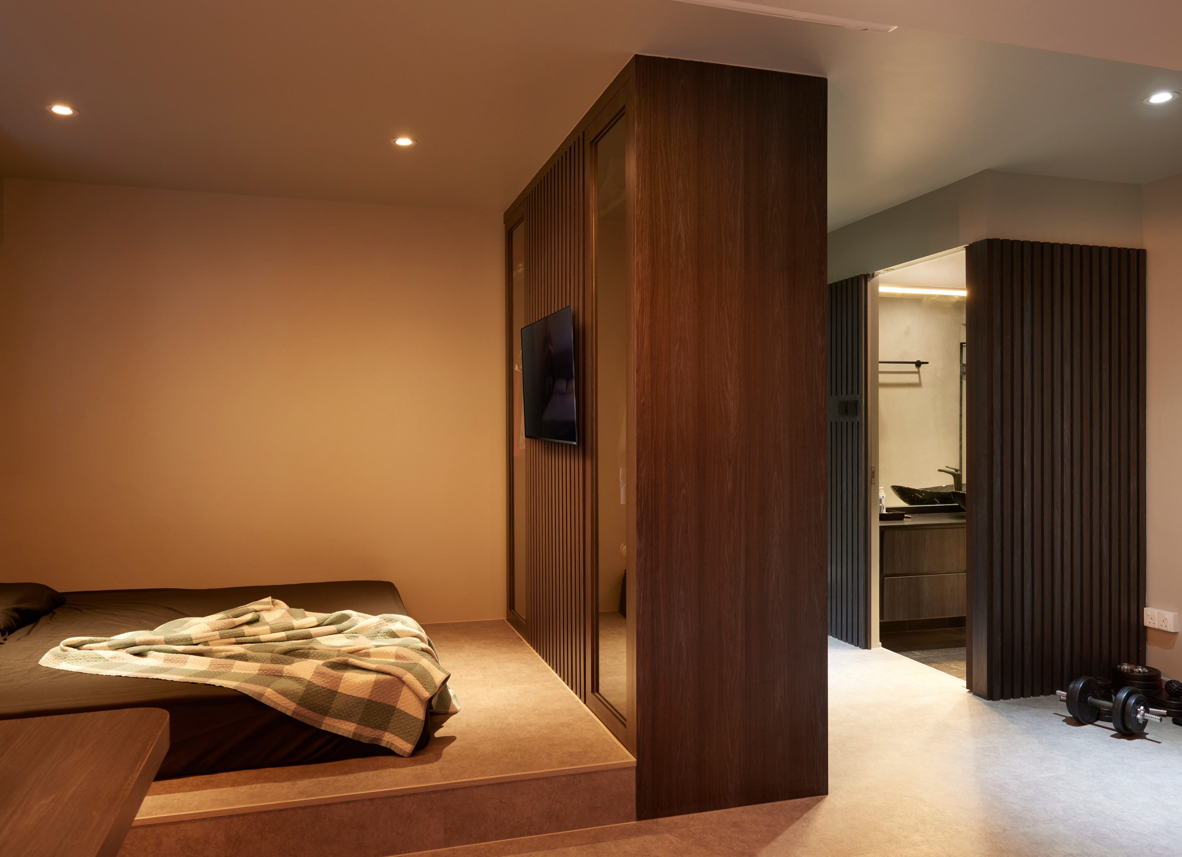 A bedroom on a raised cork-toned platform beside a walnut-veneered partition, a wall-mounted screen on fluted panelling, and a fluted timber column screening the ensuite beyond.