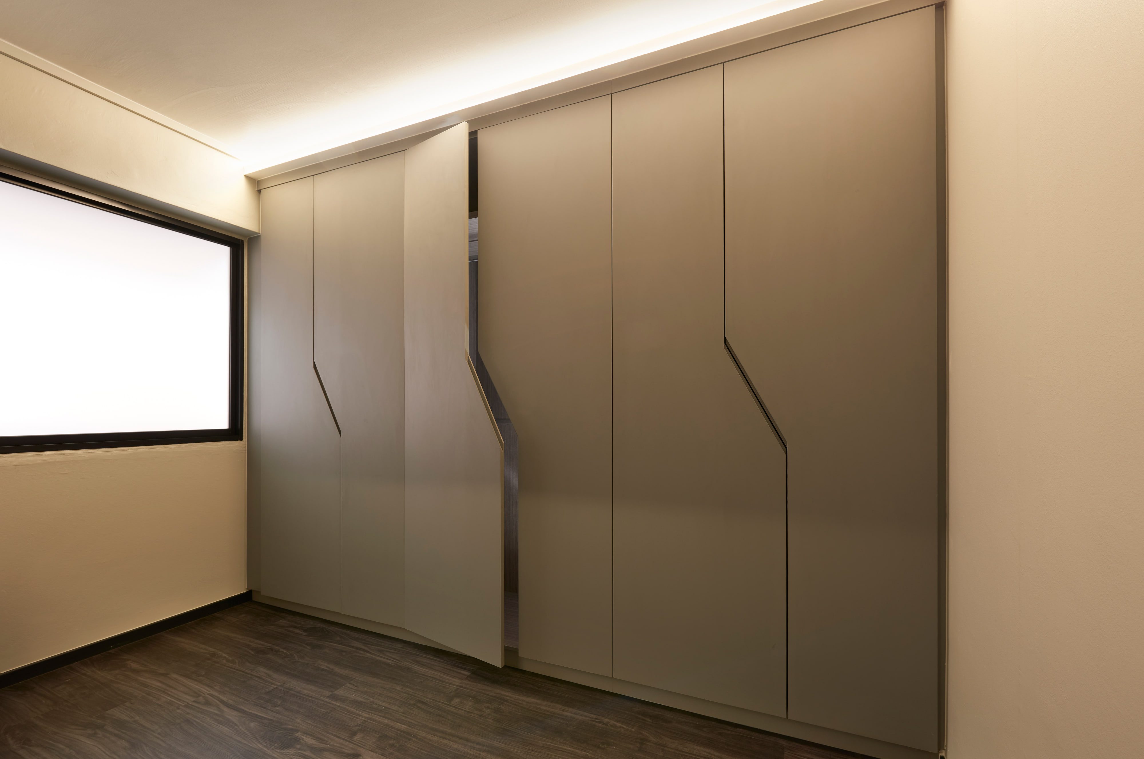 A wardrobe wall of handleless taupe doors, each cut with a single angled recess for a grip, under a continuous cove light beside a frosted window.