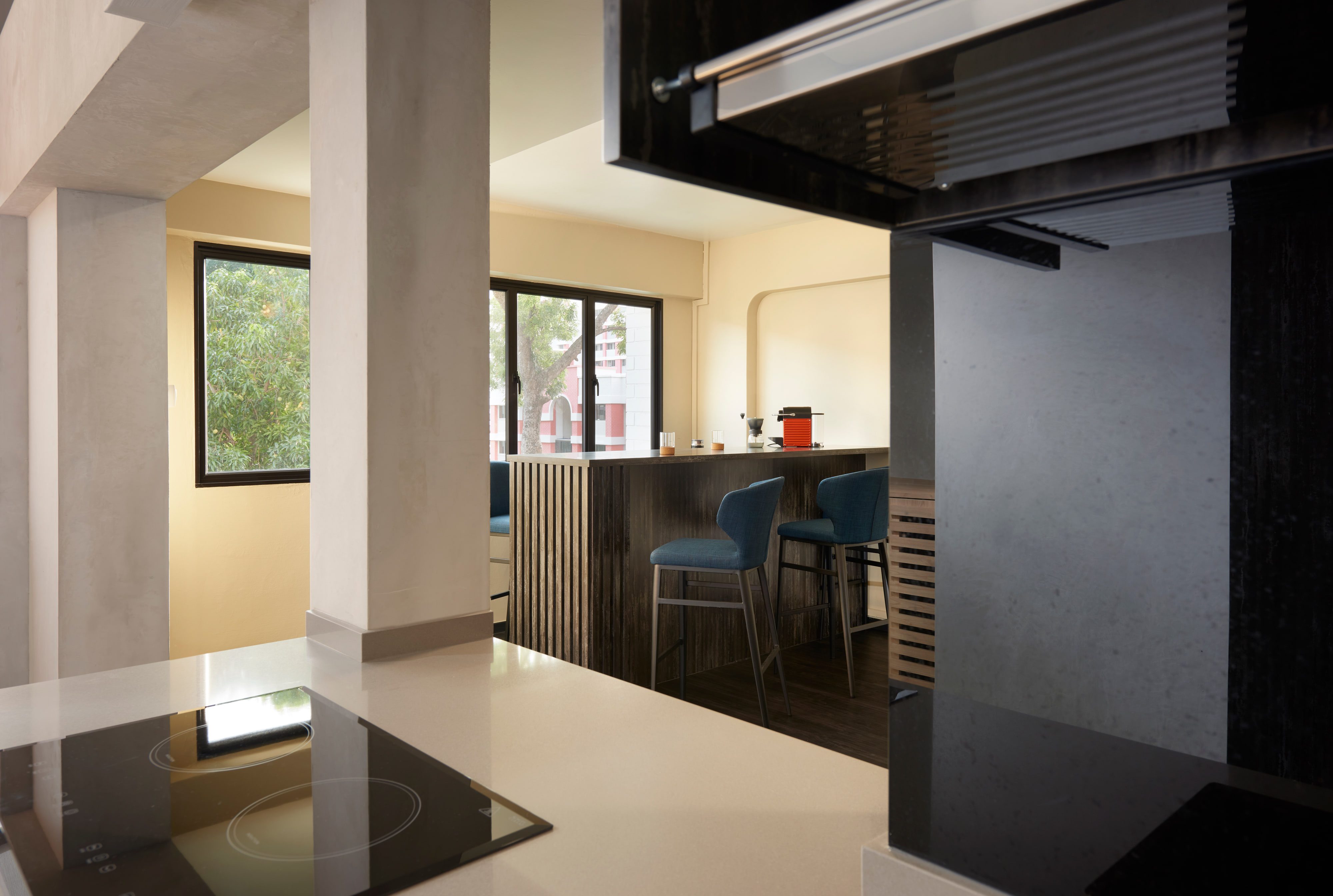 A view past a stone-clad column and an induction hob to a breakfast counter in dark timber with two teal upholstered stools, daylight and tree canopy filling the window behind.