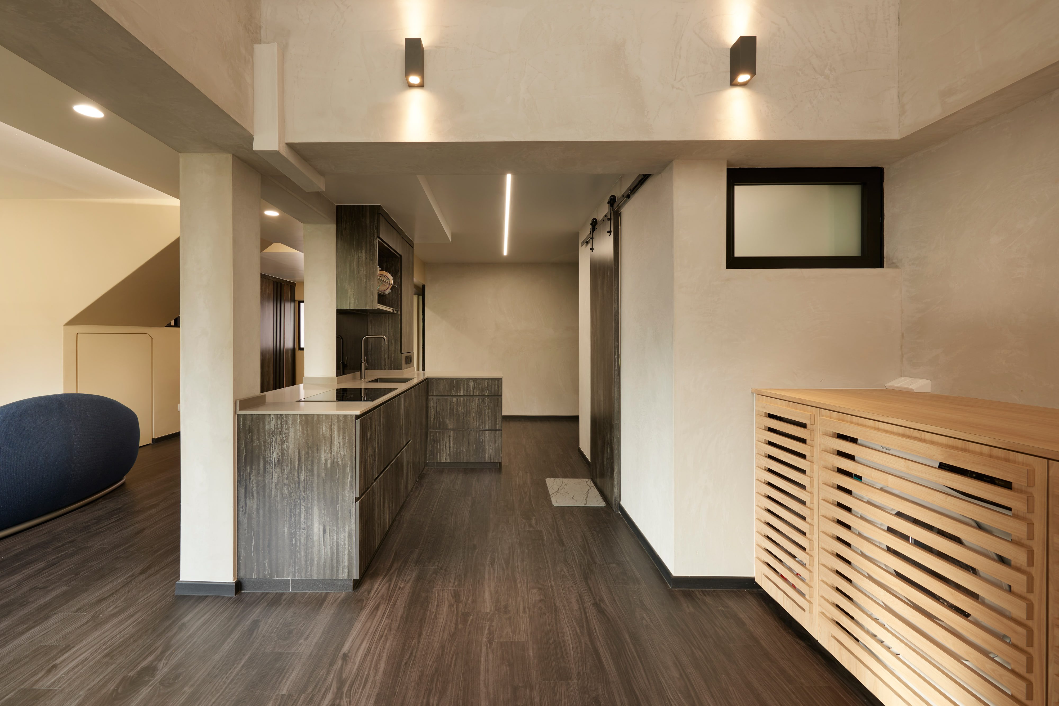 A galley kitchen in weathered grey-brown timber under a pale stone worktop, with a linear ceiling light, a sliding barn door at the far end and a slatted oak appliance cabinet in the foreground.