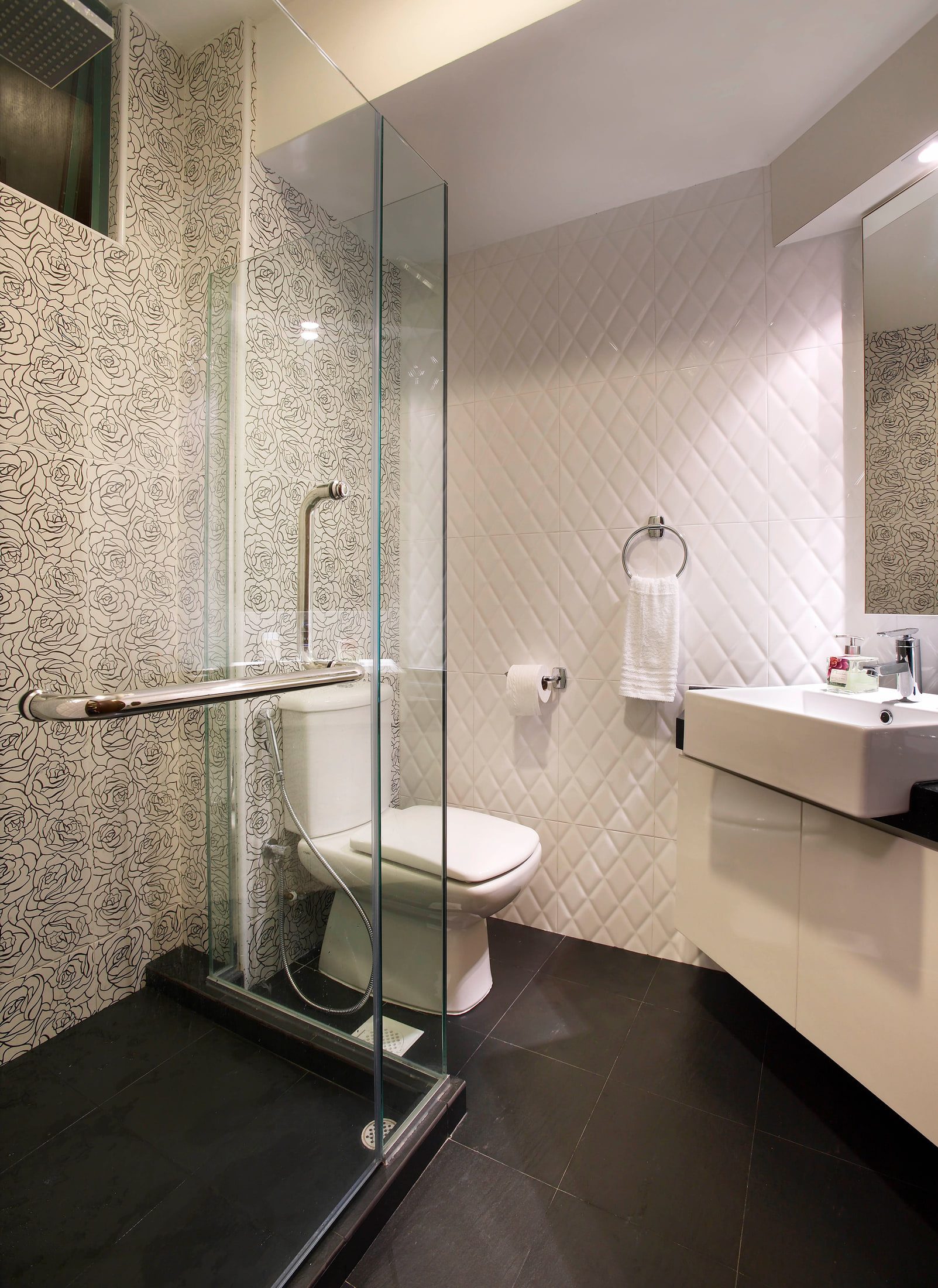 A bathroom with outlined rose pattern tile to the shower walls, a walk-in shower behind a frameless glass screen, a wall-hung WC against quilted white tile and a square vessel basin on a cream gloss vanity.