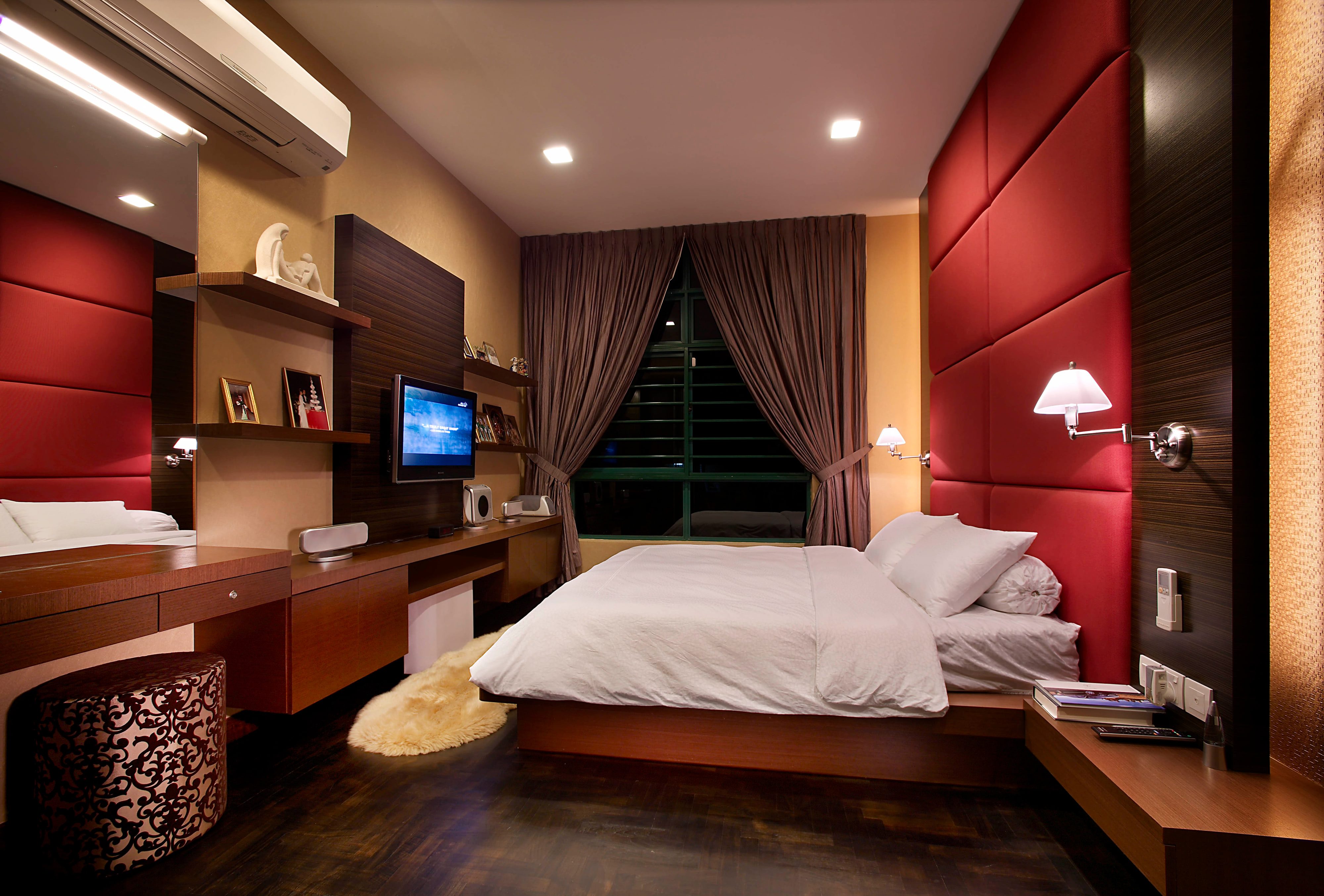 A bedroom with padded red leather headboard panels in a wide grid, chrome swing-arm wall lights either side, and a long timber desk run with floating shelves and a wall-mounted screen opposite.