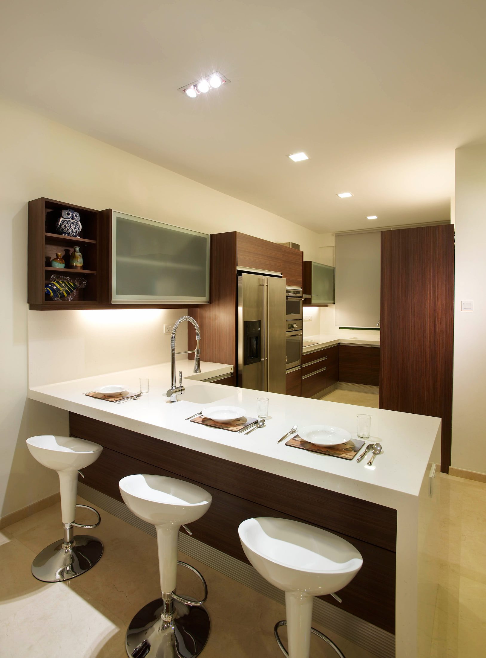 A kitchen peninsula in dark timber under a white worktop with three moulded bar stools, frosted glass wall cabinets behind it and tall dark timber units carried to the ceiling.