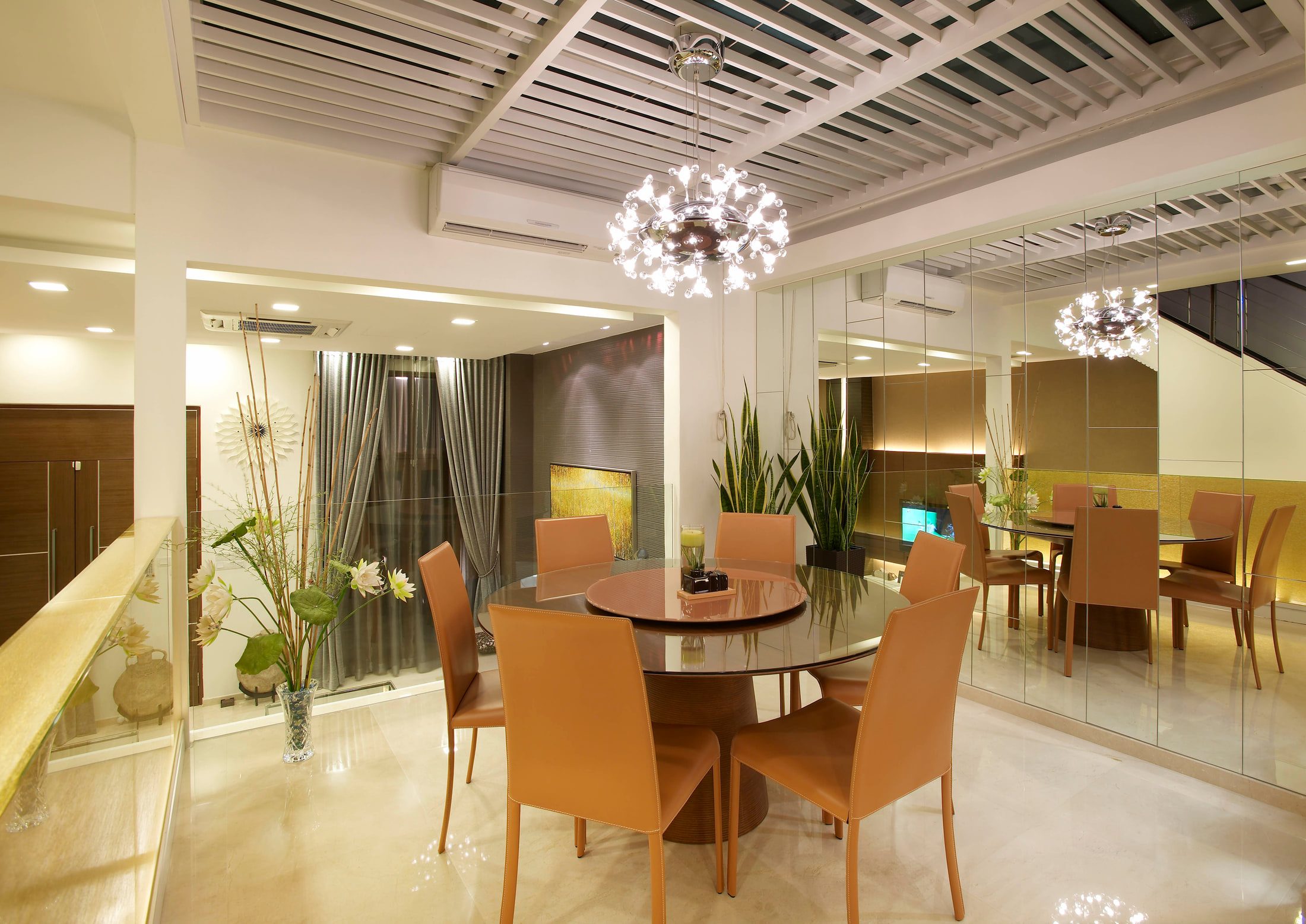 A dining room with a round dark glass table on a timber drum base, six tan leather chairs, a chrome globe chandelier under a slatted ceiling, and a full-height mirrored wall doubling the room.