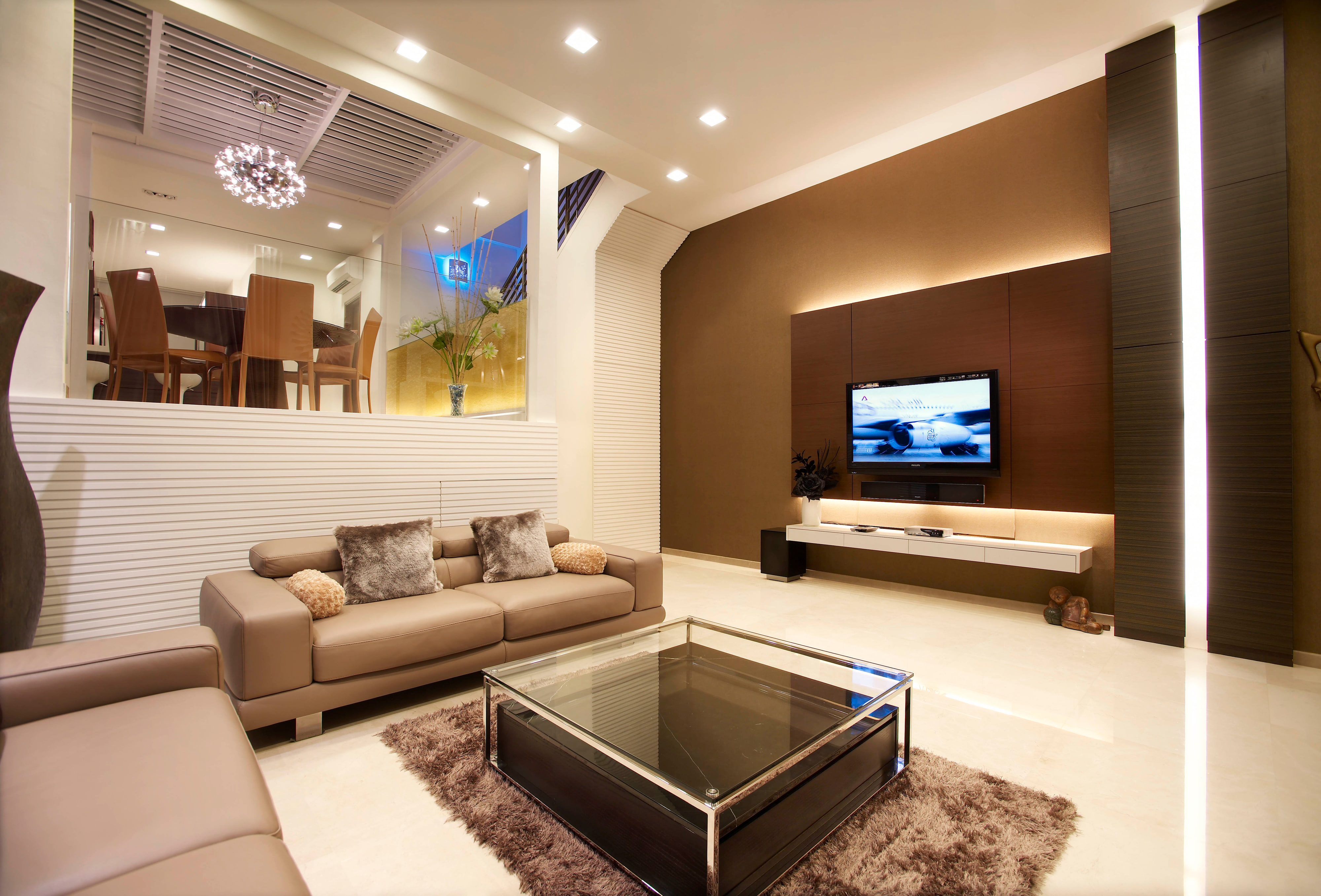 A living room with two taupe leather sofas around a glass and chrome coffee table, a flat screen on a brown panelled wall above a lit floating console, and the raised dining room visible beyond a slatted white half-wall.