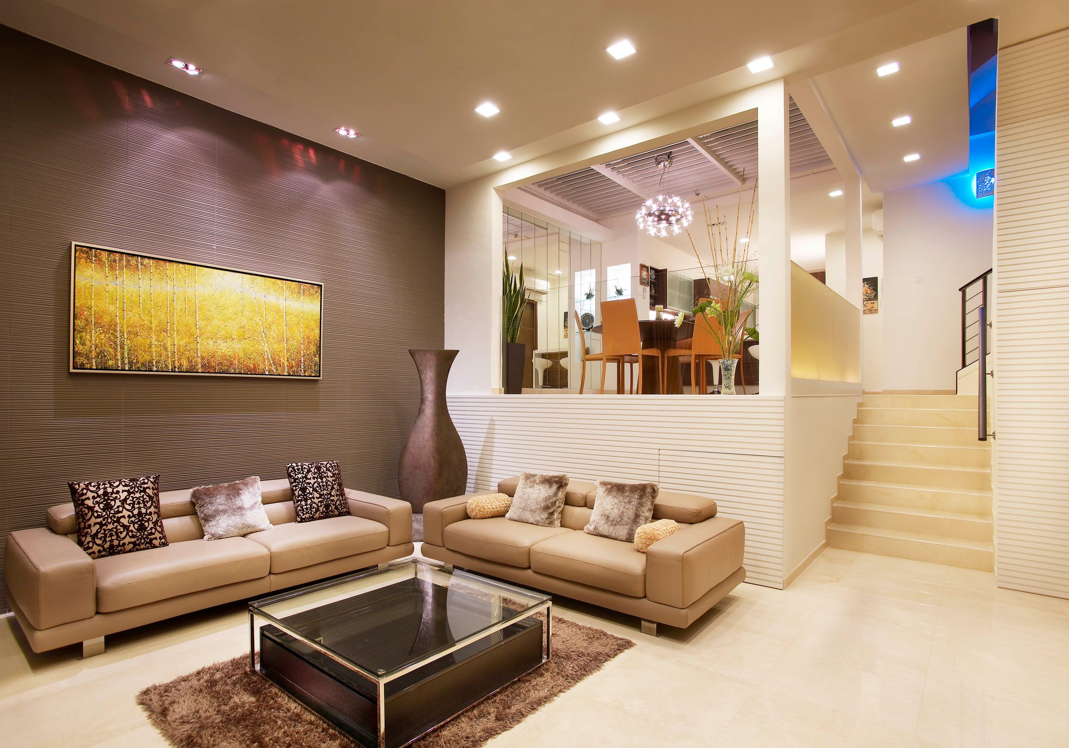 A living room seen from the stair end, with a wide framed landscape of golden birches on a brown textured wall, two taupe leather sofas around a glass coffee table, and a pale stone staircase rising behind a black steel handrail.