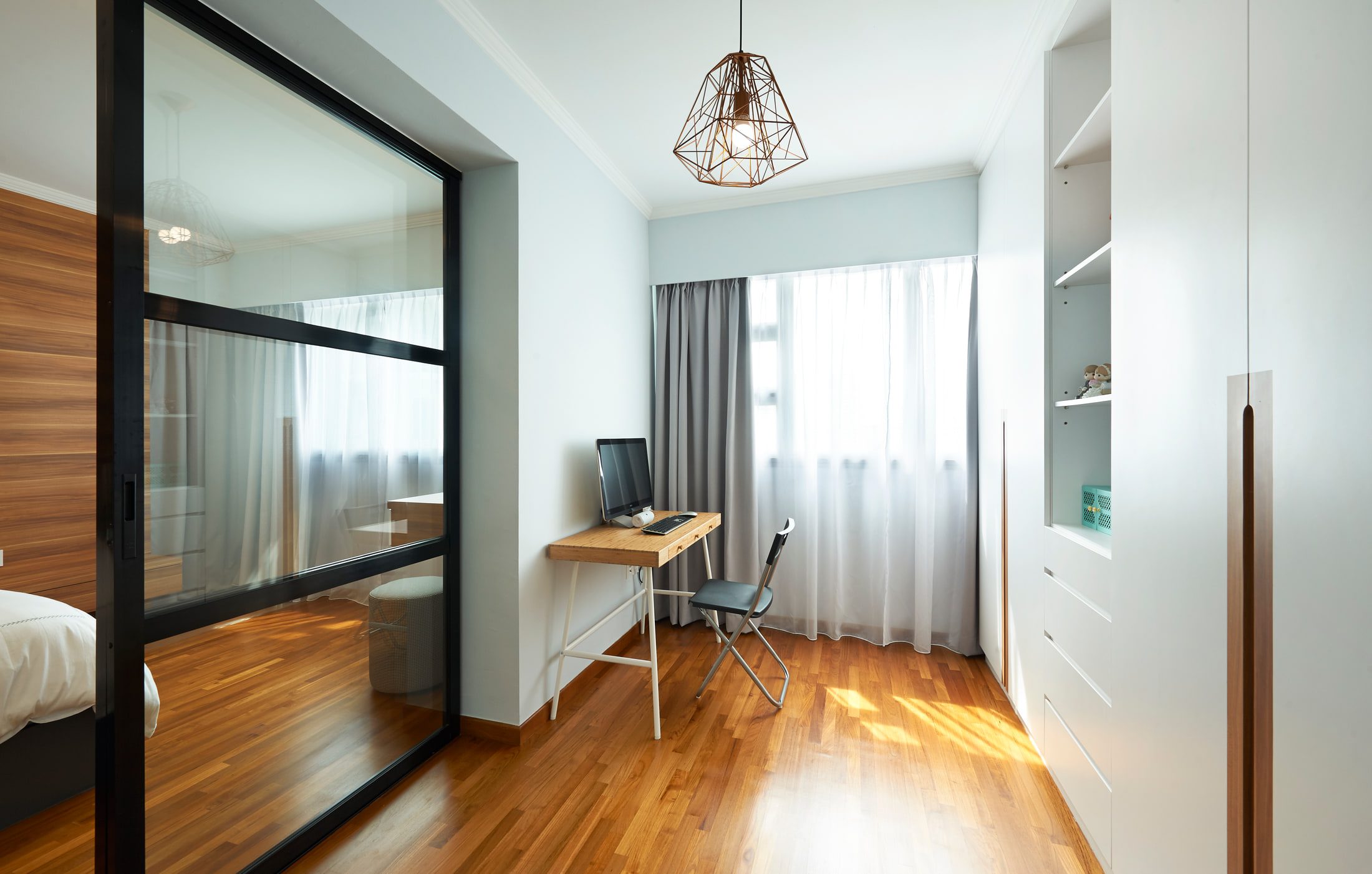 The study seen through the bedroom's black-framed glazed screen, a bamboo desk and folding chair set under a copper wire pendant on strip-parquet flooring.