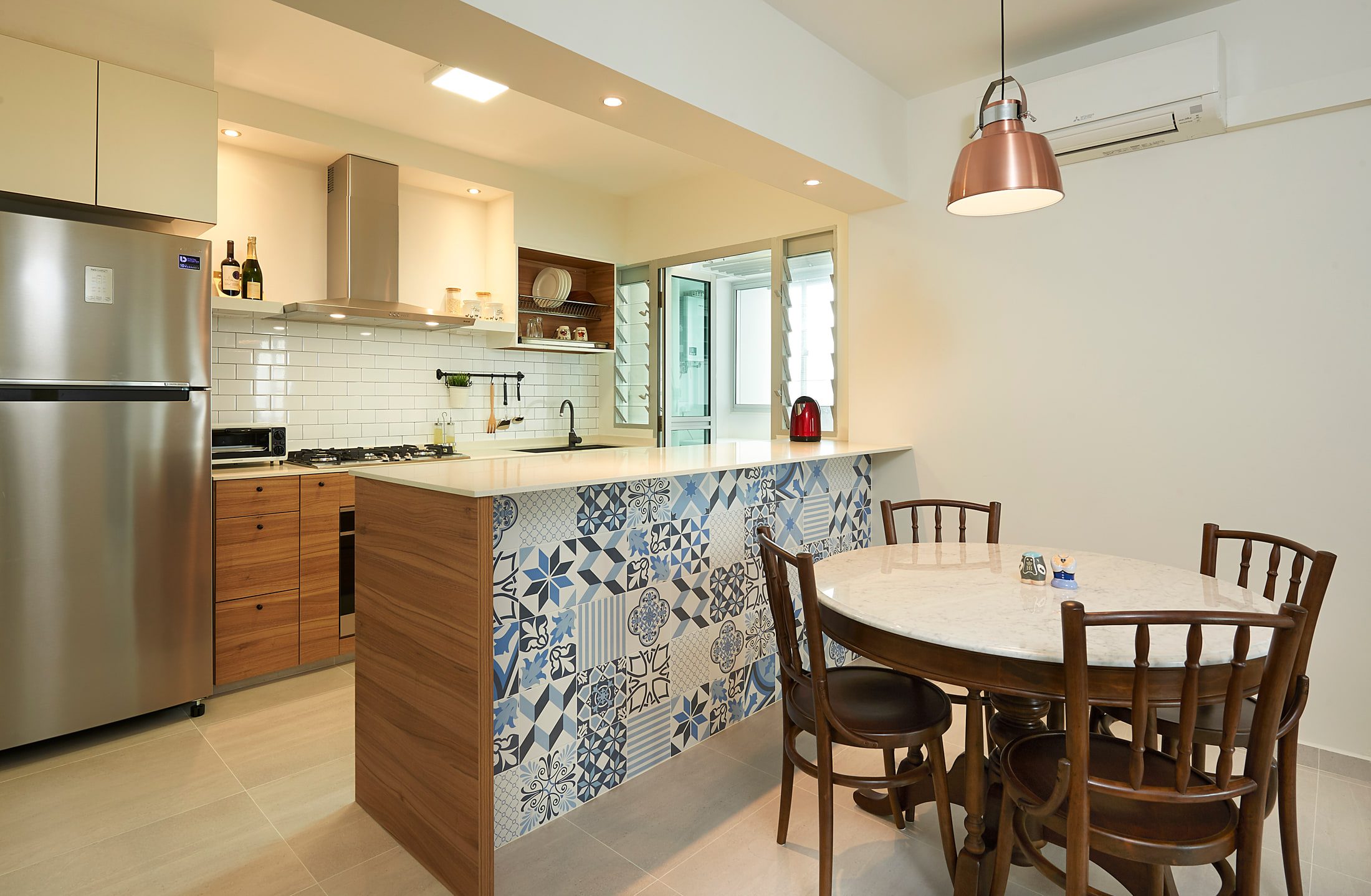 An open kitchen and dining area where a white quartz peninsula faced in blue and white patterned cement tile meets a round marble table and dark bentwood chairs.