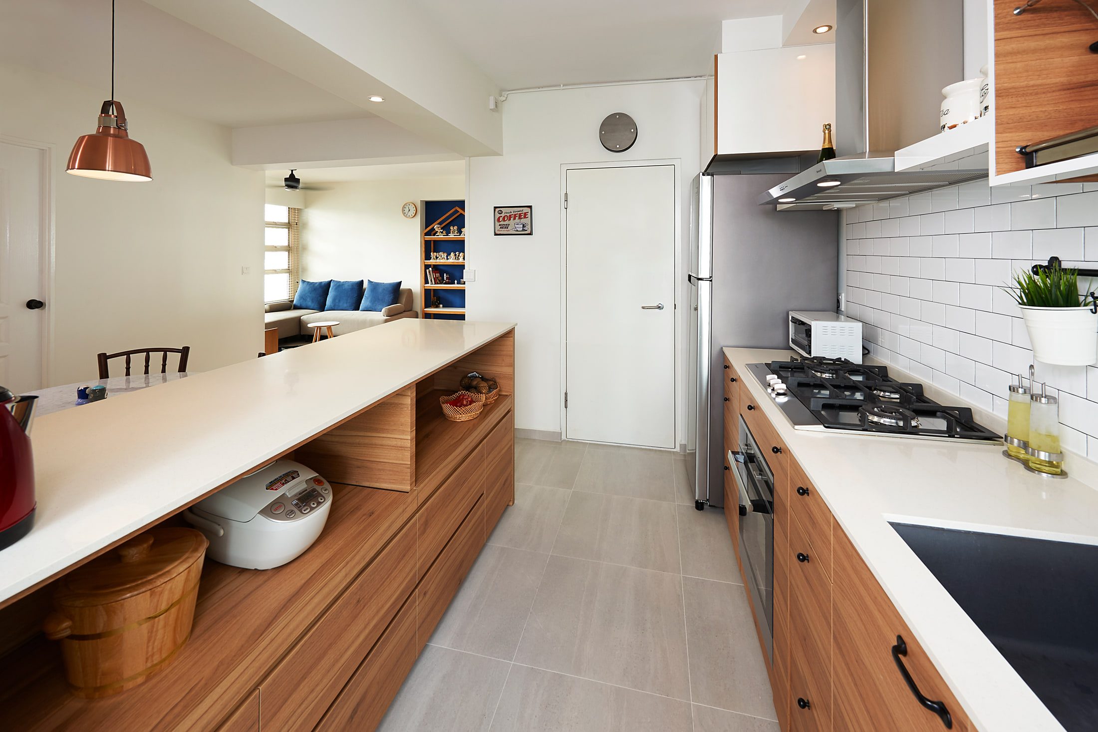 The galley kitchen looking through to the living room, a long white worktop with open timber cubbies below it and a copper dome pendant over the breakfast counter.