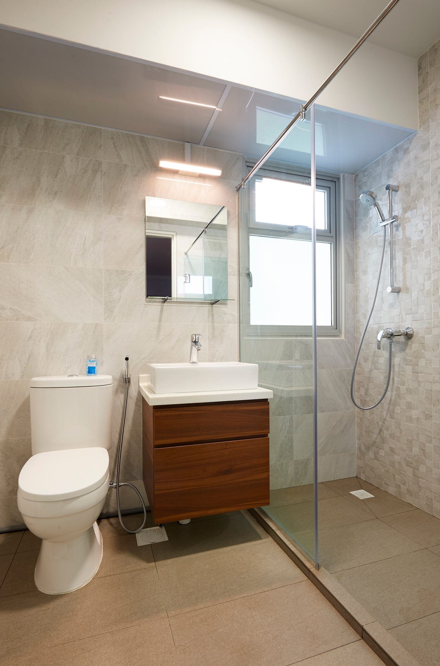 A bathroom lined in pale stone-effect tile with a walnut wall-hung vanity, a white rectangular vessel basin and a frameless glass screen dividing off the shower.