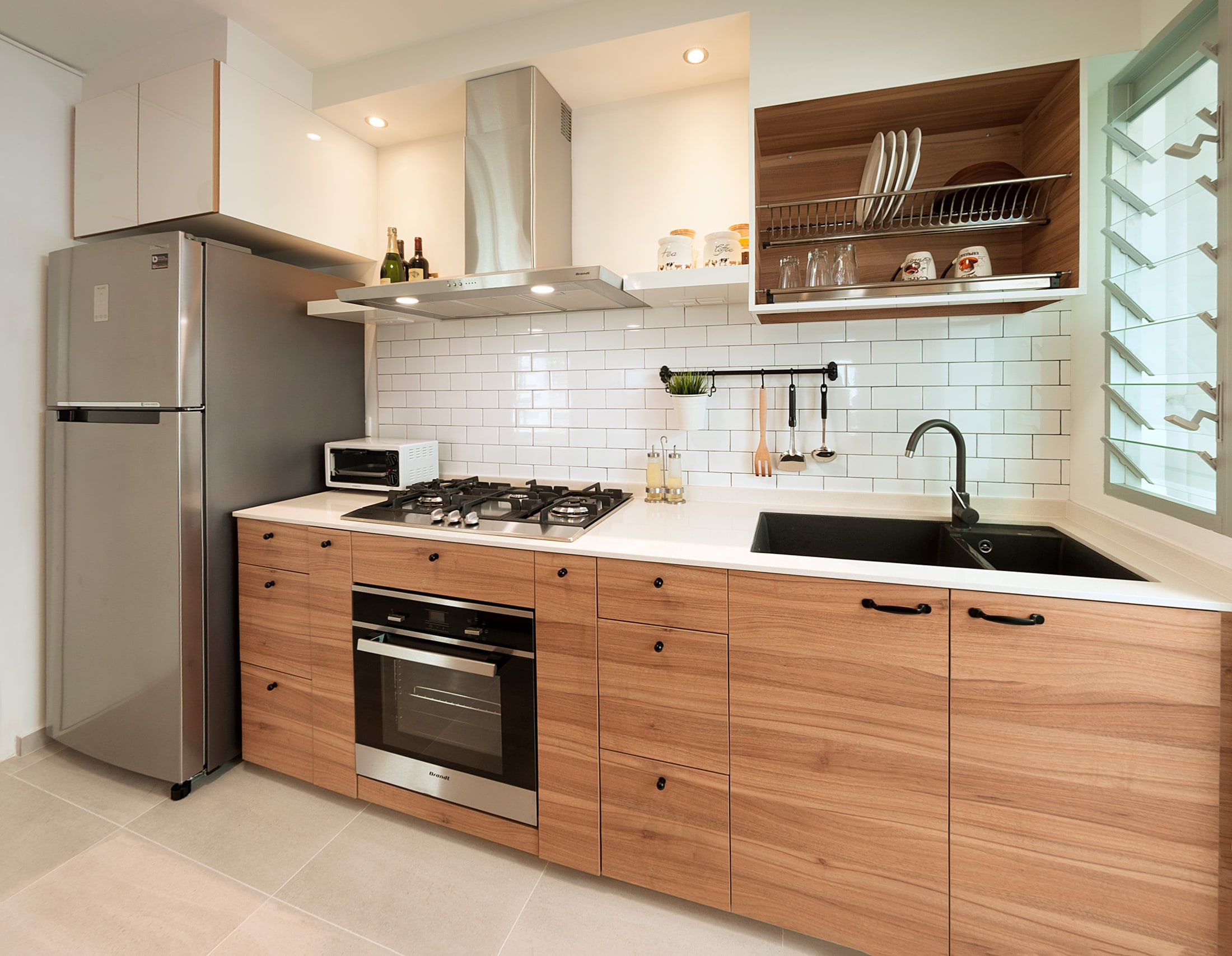 A kitchen run in walnut-grain fronts under a white quartz worktop, with a white metro-tile splashback, a stainless steel hood and a timber dish-rack cabinet beside a louvred window.