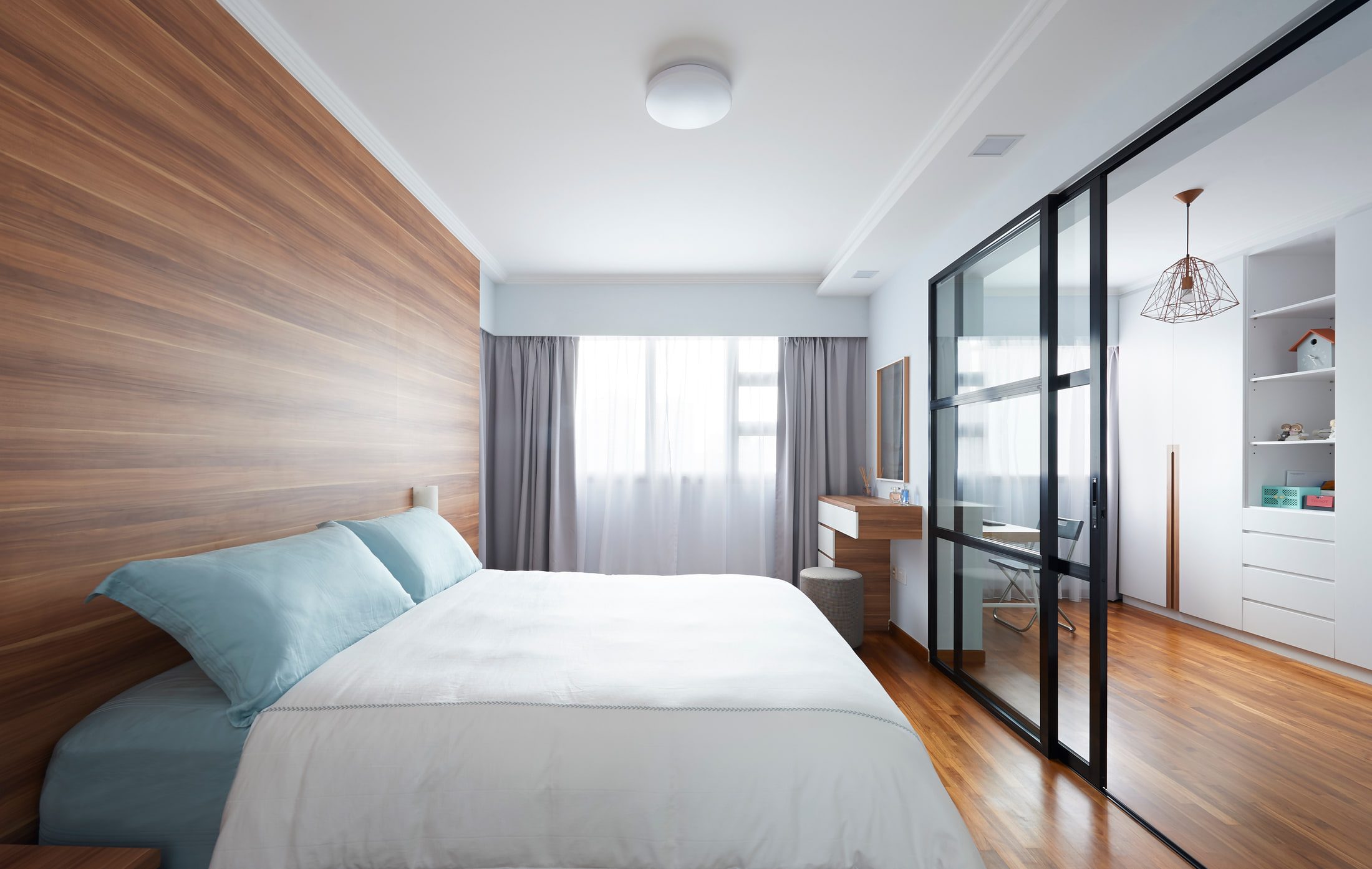 A bedroom with a walnut-panelled headboard wall, blue and white bedding, and a black-framed glazed partition opening to a study.