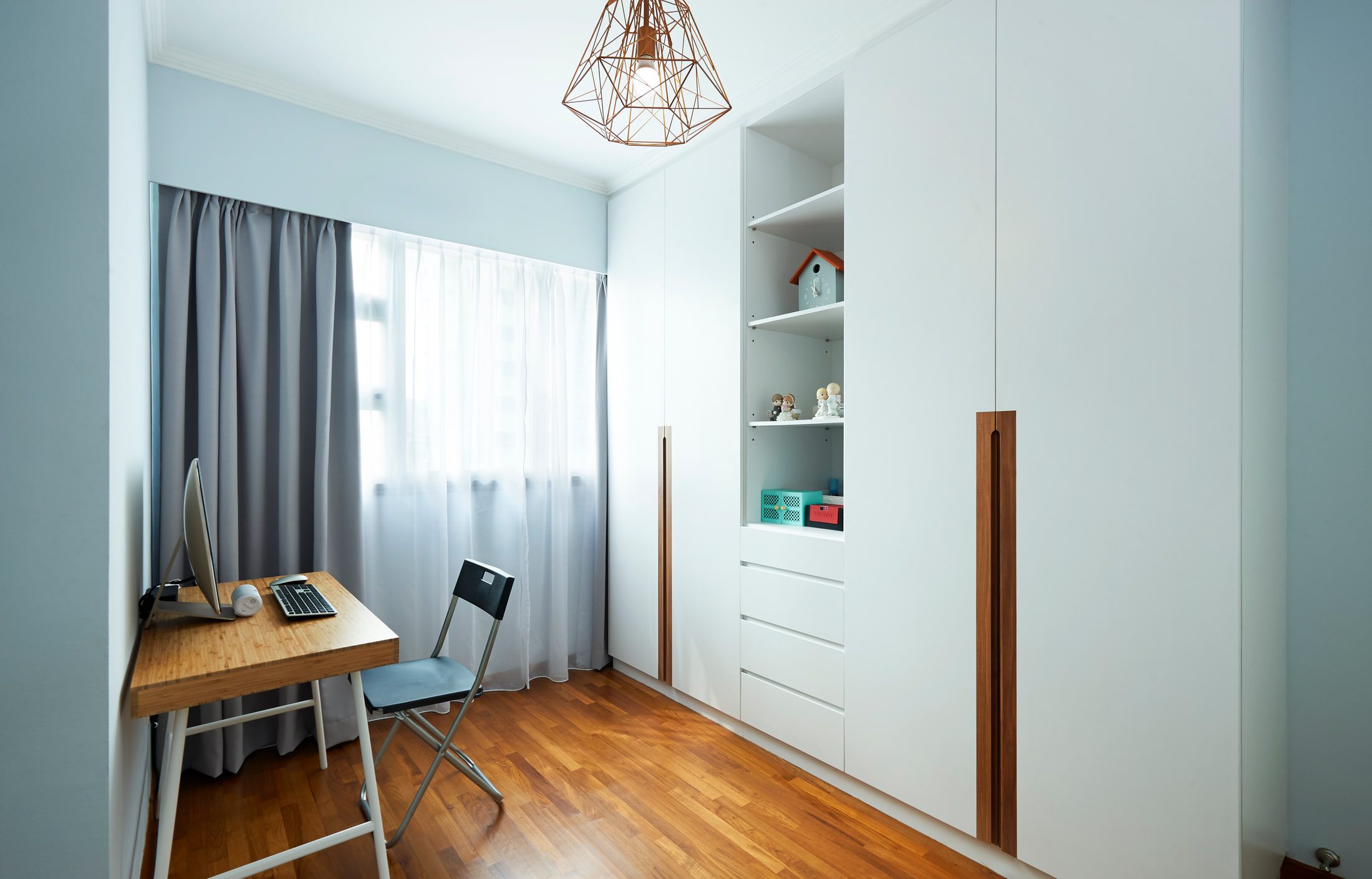 A narrow study lined with full-height white wardrobes with routed timber pulls, an open shelved bay of ornaments, and a bamboo desk against the curtained window.